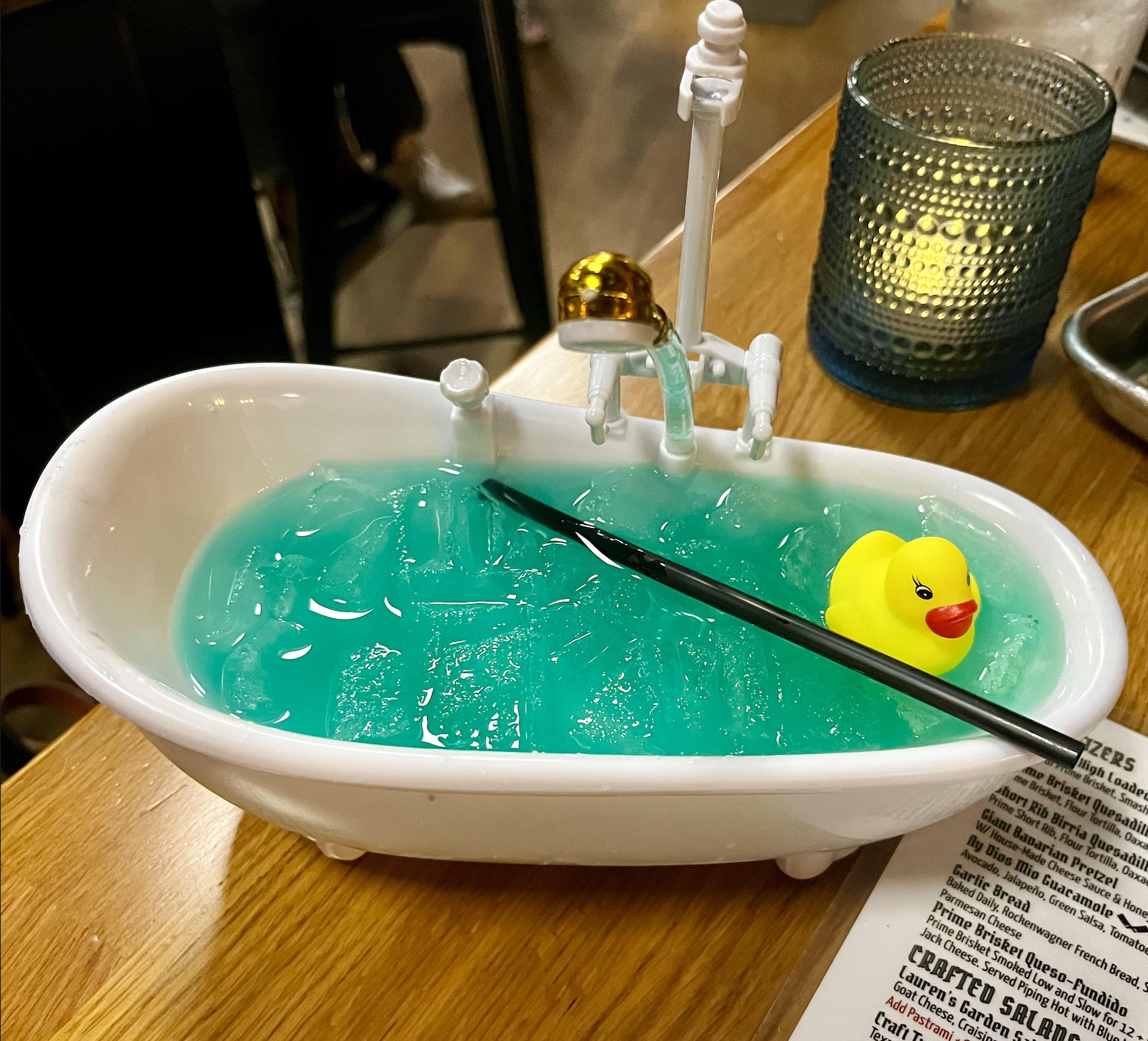 A blue cocktail with ice is served in a small bathtub-shaped container, garnished with a mini yellow rubber duck and a black straw. A lit candle and menu are nearby on the wooden table.