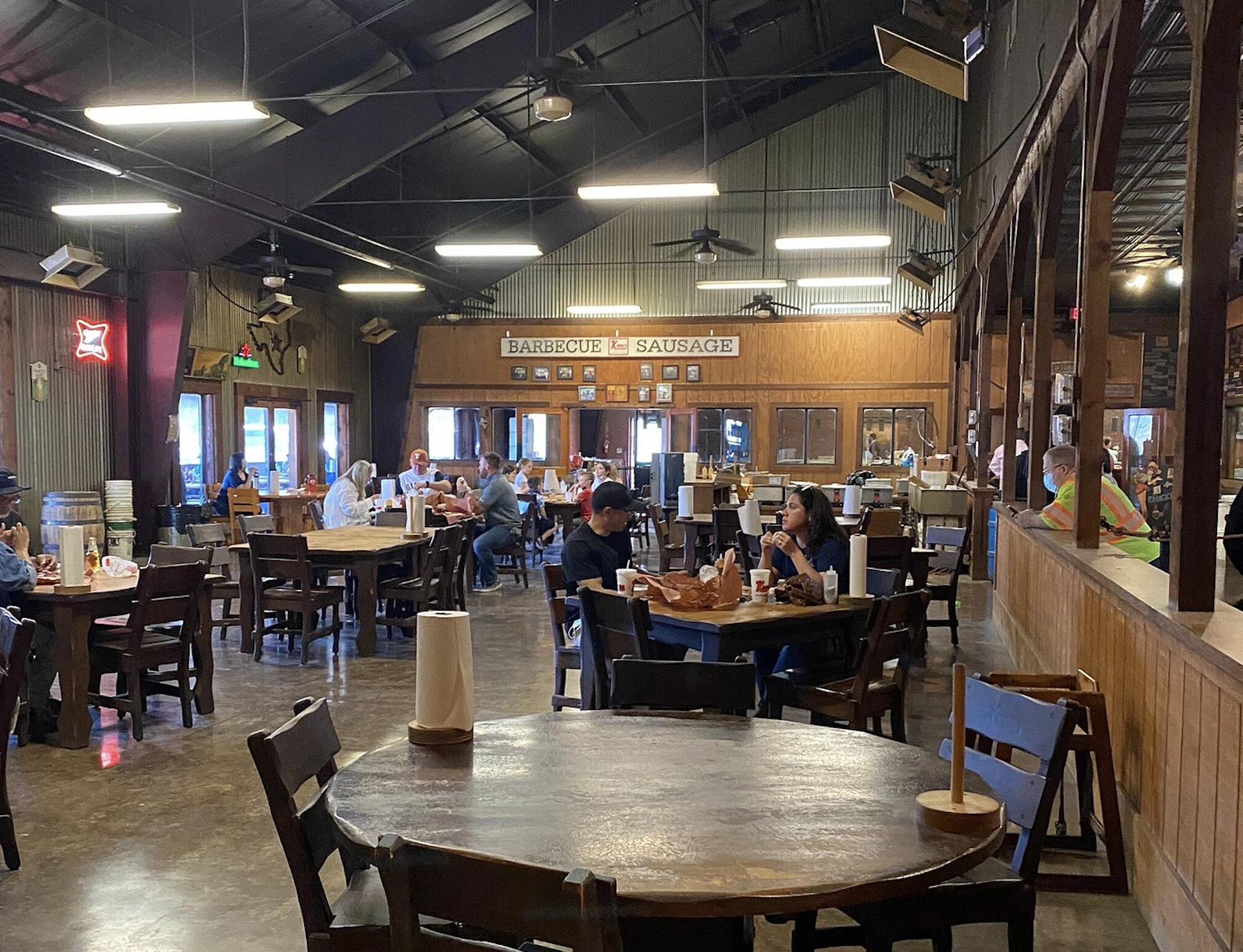 People sit at wooden tables inside a rustic barbecue restaurant with brown walls and high ceilings. A “Barbecue Sausage” sign hangs on the far wall. Paper towels are on each table.