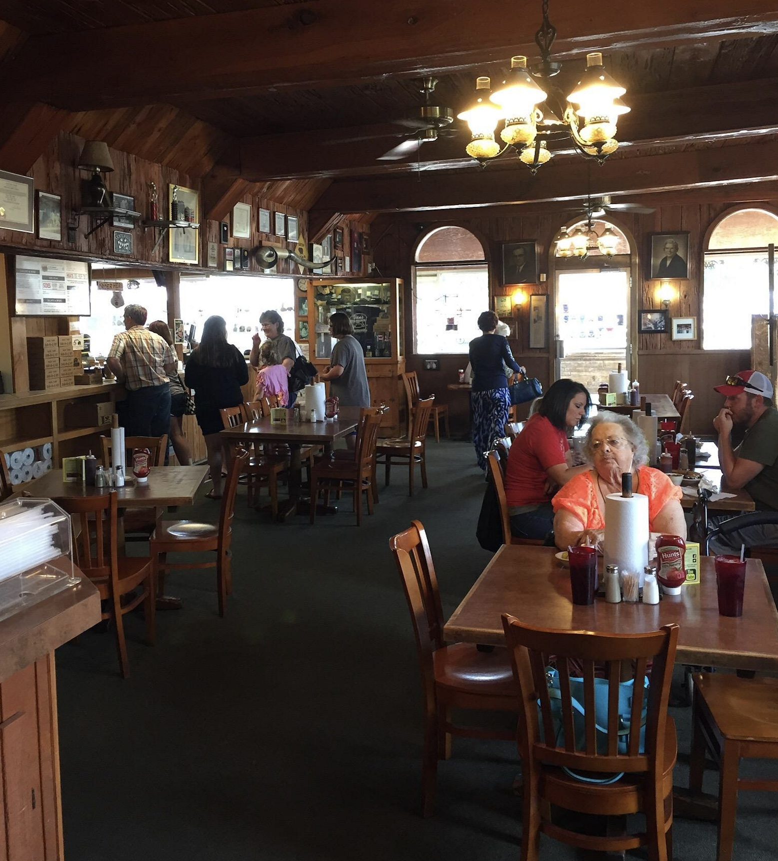 Casual restaurant interior with wooden walls and ceiling, several people sitting at tables, others standing at the counter. Daylight streams in through arched windows, creating a warm, inviting atmosphere.