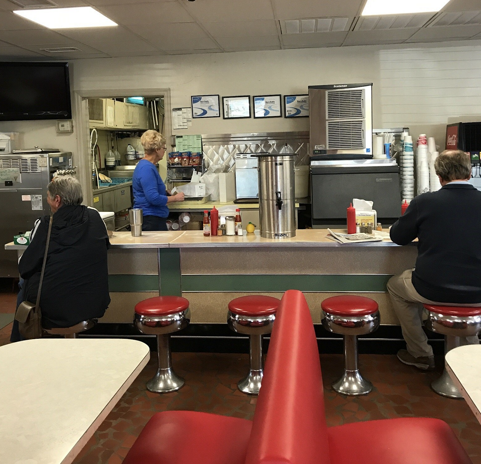 A classic diner with red stools and booths. Three people are seated at the counter, while a woman stands behind it. The counter has condiments and a large coffee dispenser. The diner interior looks retro and cozy.