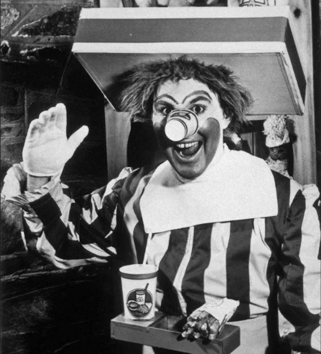 A black-and-white photo of a person dressed as a clown with a striped outfit, large bow tie, and exaggerated makeup, holding a cup and waving while appearing to pop out of a box.