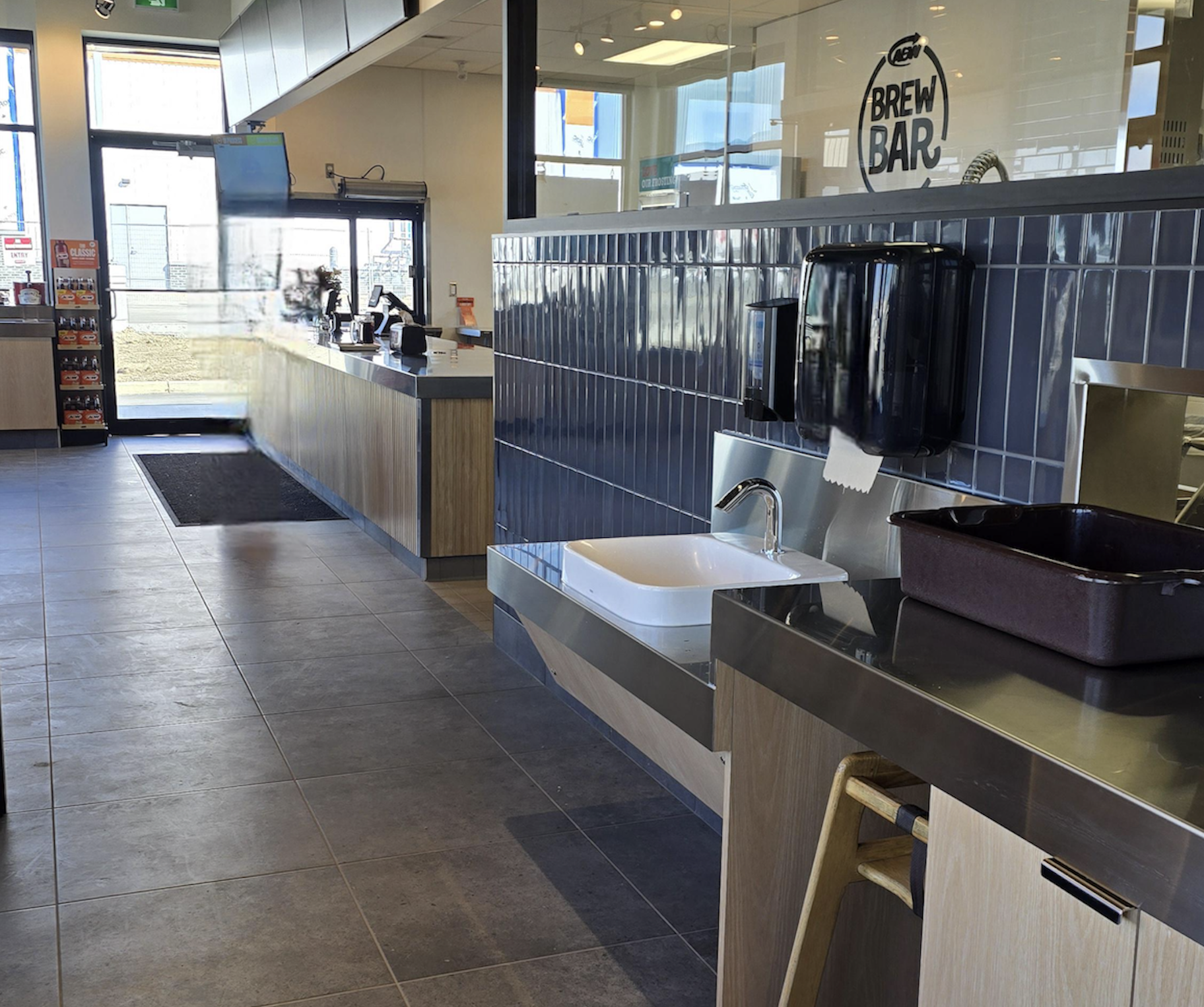 Modern self-serve coffee station with a sink, paper towels, coffee machines, and clean tiled walls. Large windows let in natural light. Floor is tiled and counters are wood-accented.