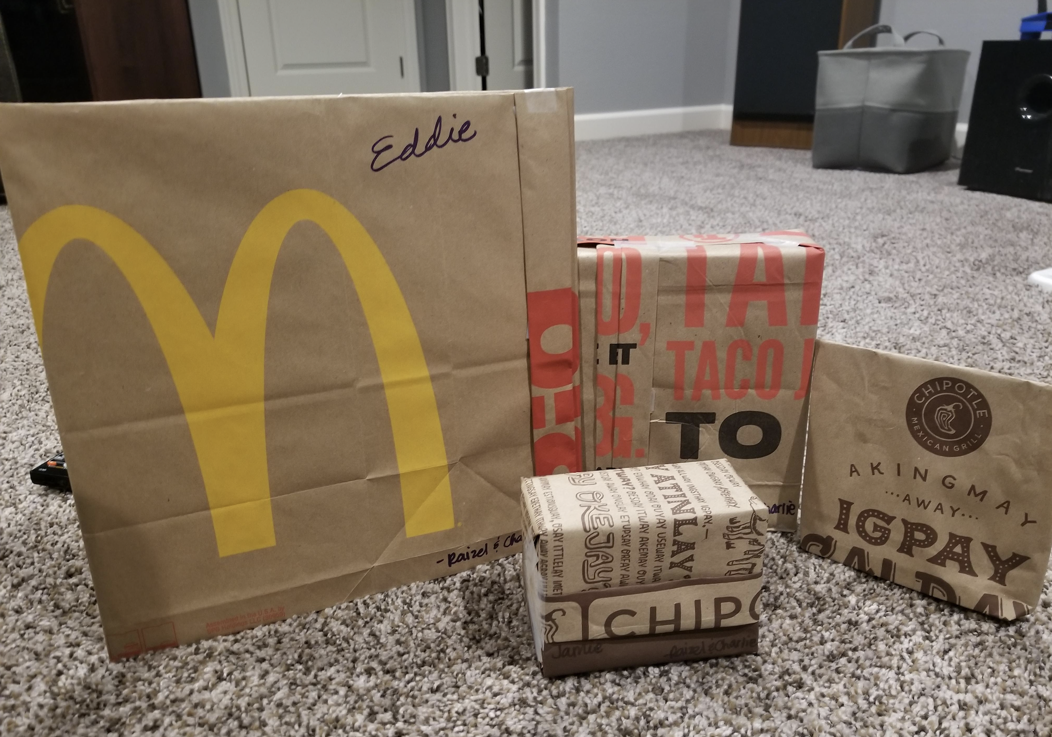 Four fast food takeout bags and boxes, including McDonald’s and Chipotle items, sit on a carpeted floor; “Eddie” is written on the McDonald’s bag.