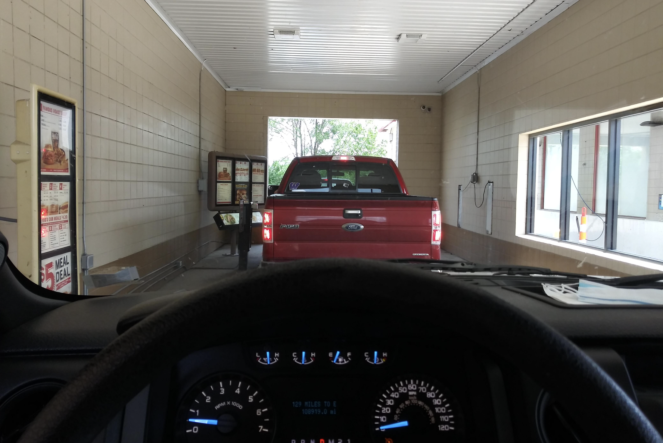 View from inside a vehicle at a drive-thru, showing a red pickup truck ahead waiting at the order speaker, with menus on the left and a window on the right. The steering wheel and dashboard are visible in the foreground.