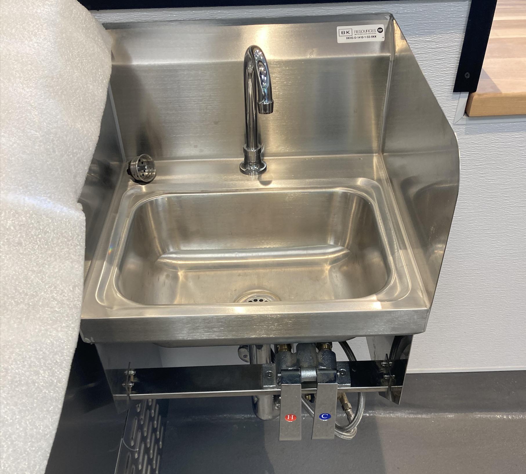 A stainless steel sink with a tall faucet, foot pedals for hot and cold water, and protective side splashes, mounted against a wall in a commercial or industrial setting.