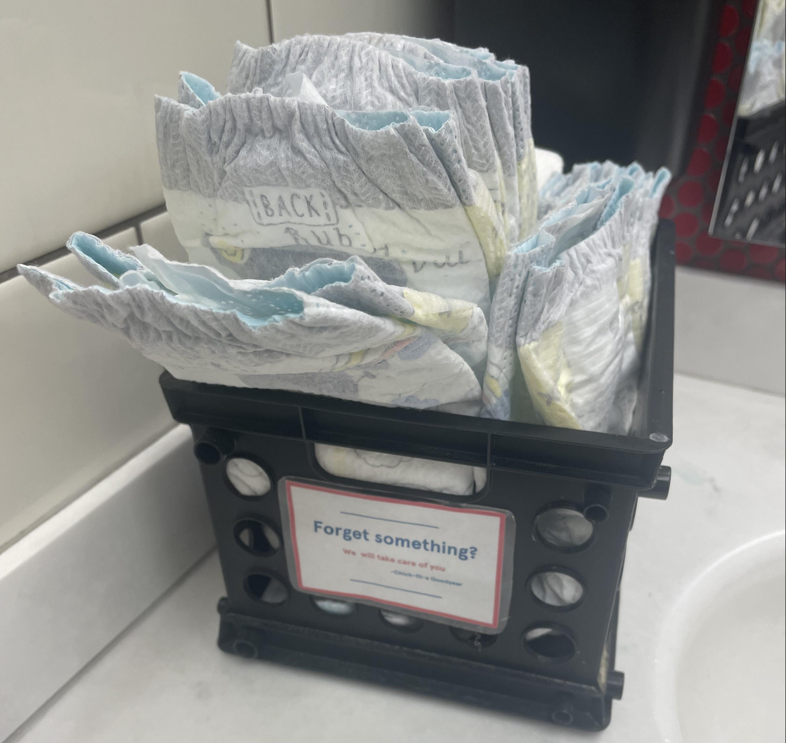 A black crate holds baby diapers in a restroom. A sign on the crate reads, “Forget something? We’ll take care of you.” The crate sits on a white countertop next to tiled walls.