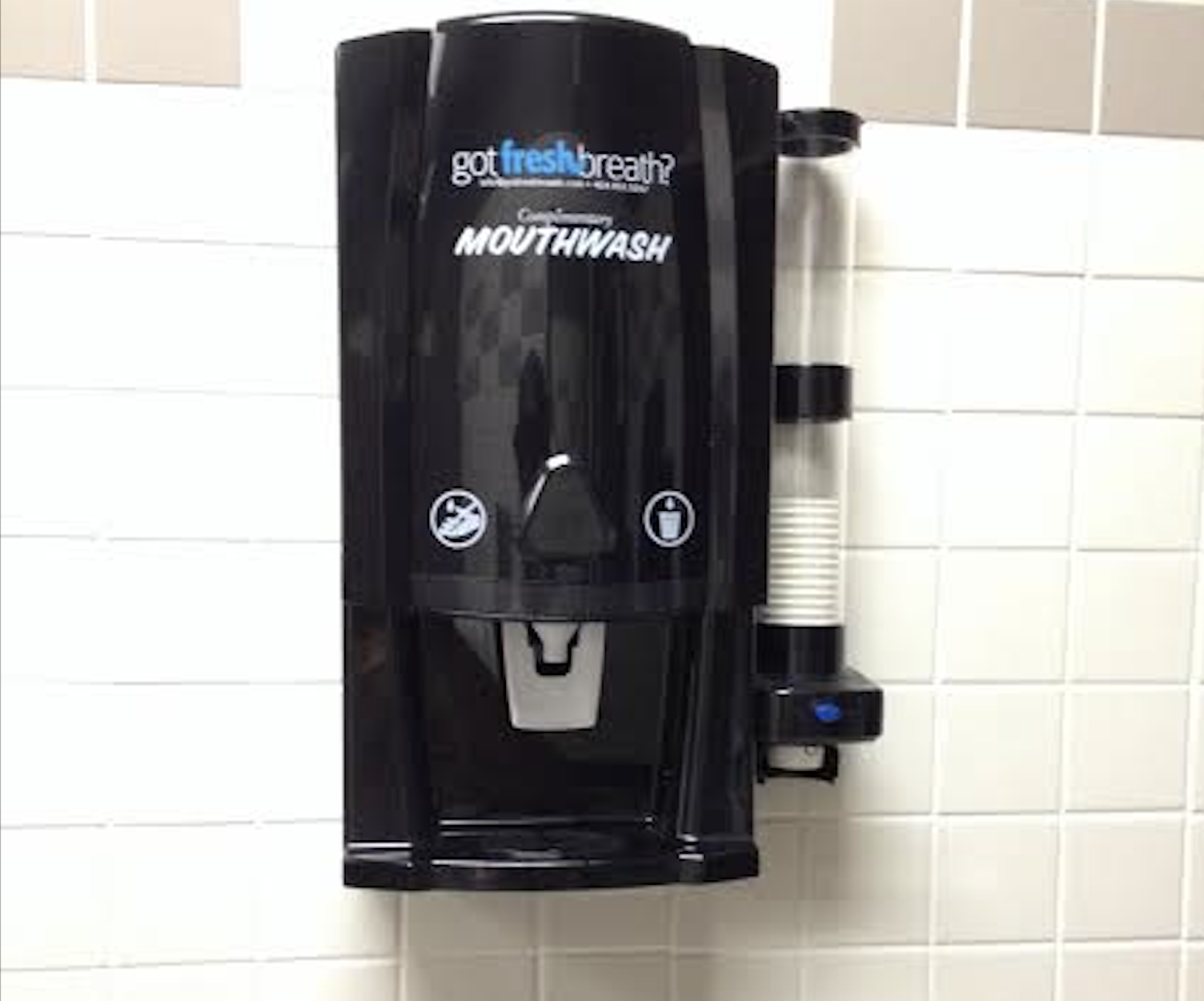 A black mouthwash dispenser is mounted on a tiled wall, with a cup holder containing stacked disposable cups attached on the right side.