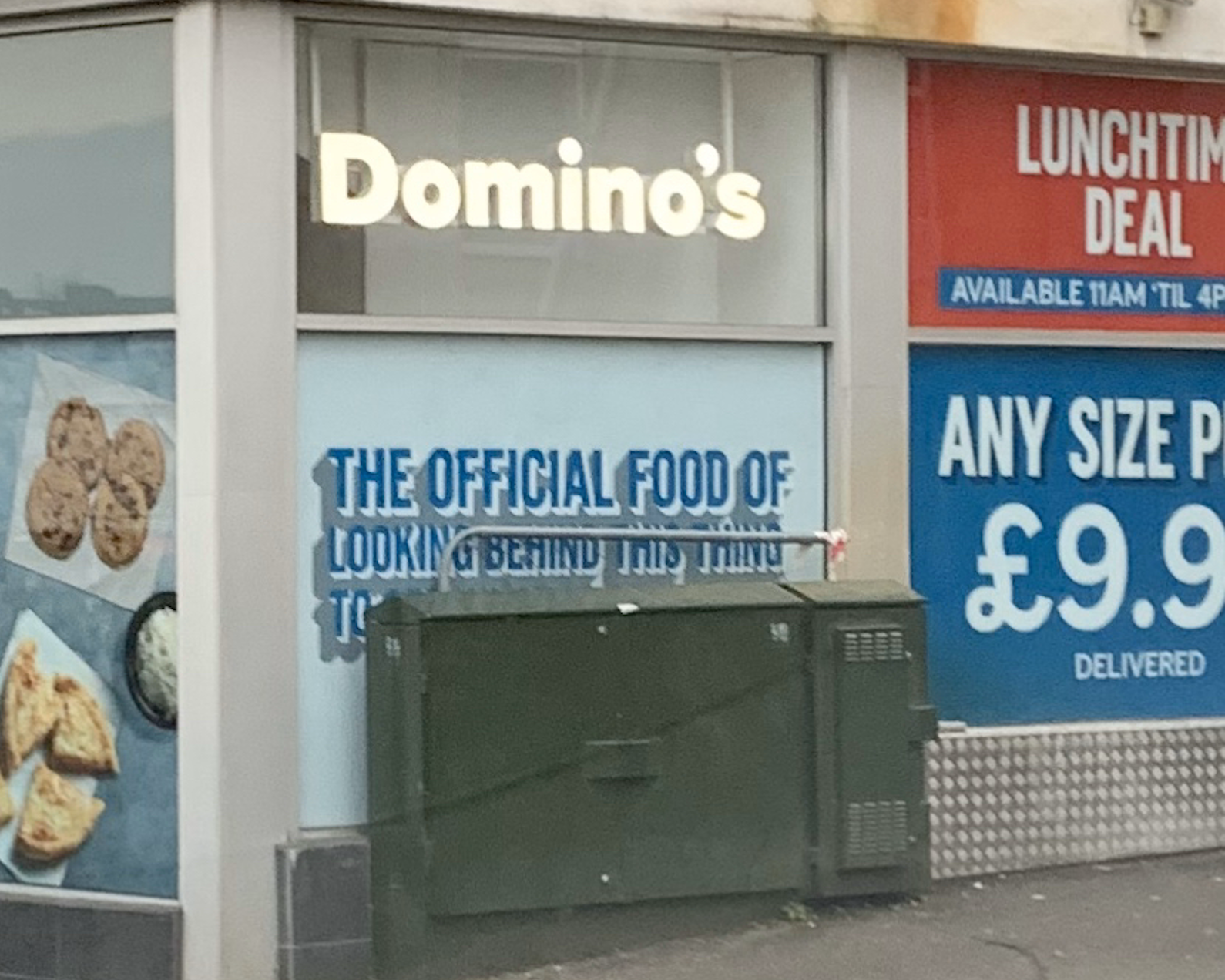 Domino’s storefront with a sign saying “THE OFFICIAL FOOD OF LOOKING BEHIND THIS THING TO READ THIS SIGN.” Part of the sign is obscured by a green utility box.