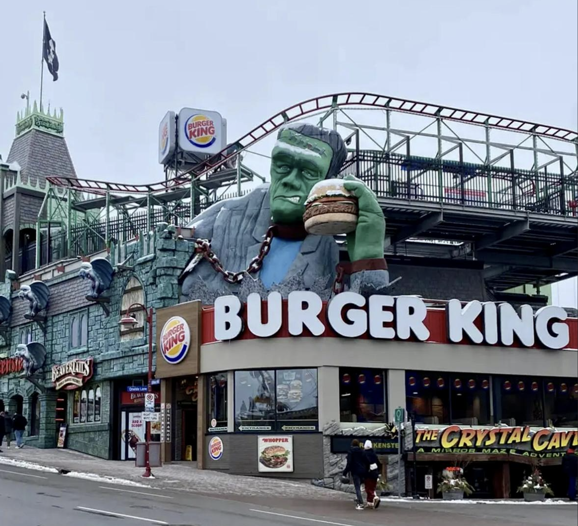 A Burger King restaurant with a giant statue of Frankenstein holding a burger on its roof, located next to a haunted house attraction, with a roller coaster track above.