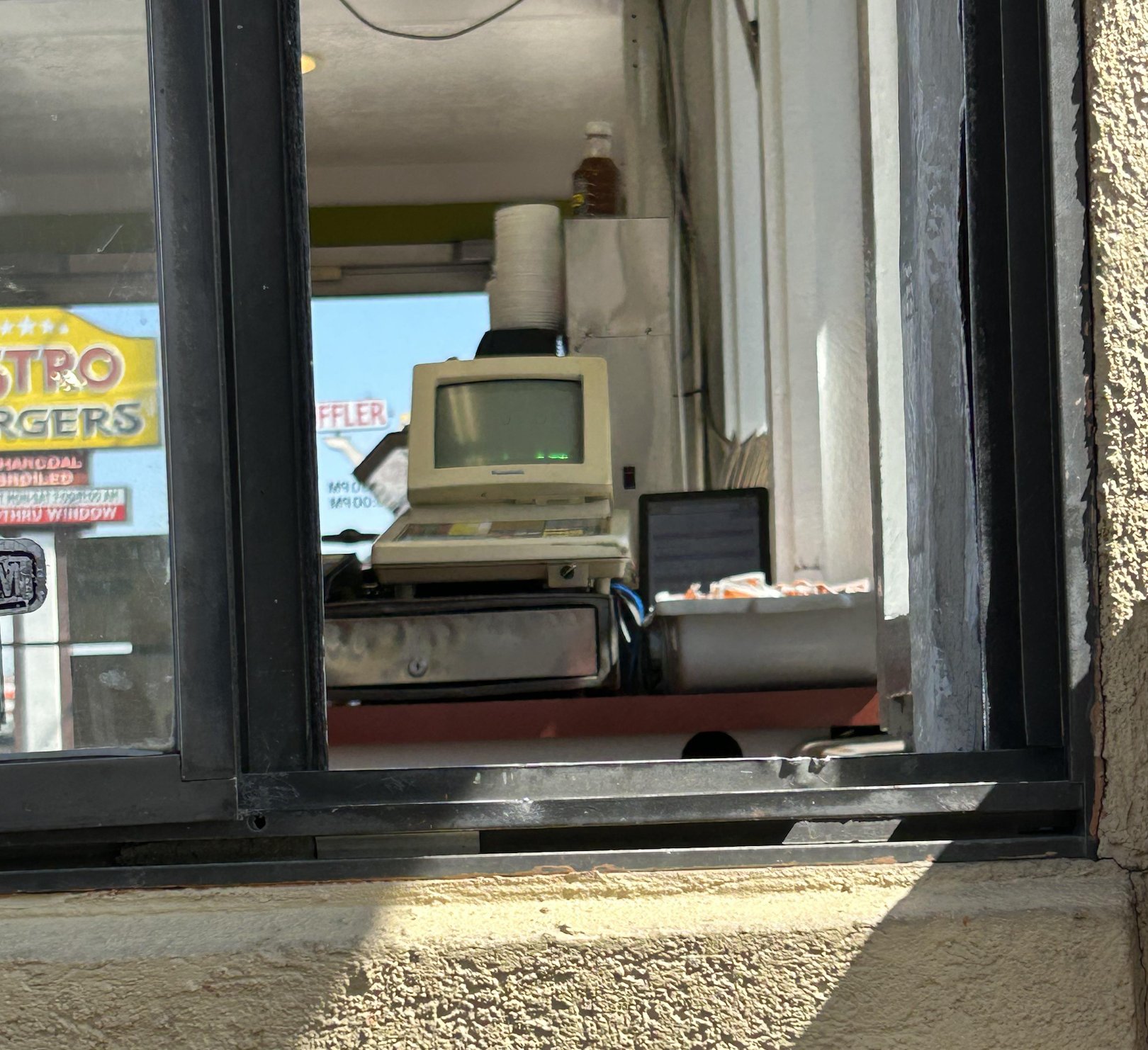 A vintage cash register sits by a drive-thru window with paper cups stacked on top. A sign for Astro Burgers is visible in the background through another window.