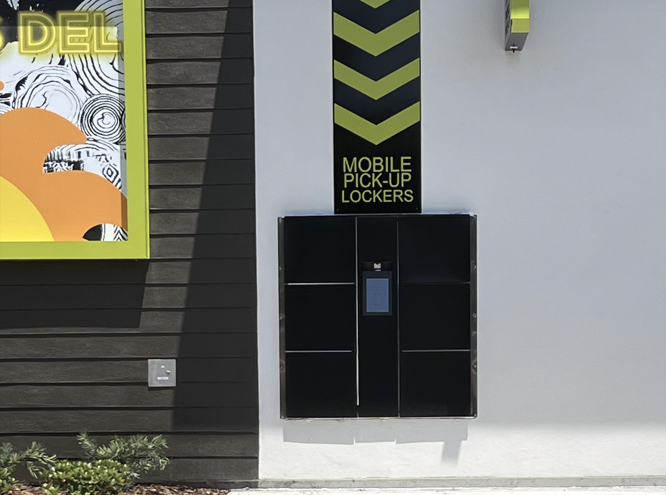 Outdoor black mobile pick-up lockers with a screen in the center, located on a white wall beneath a sign reading “MOBILE PICK-UP LOCKERS” and a large chevron pointing downward. Part of a colorful mural is visible to the left.