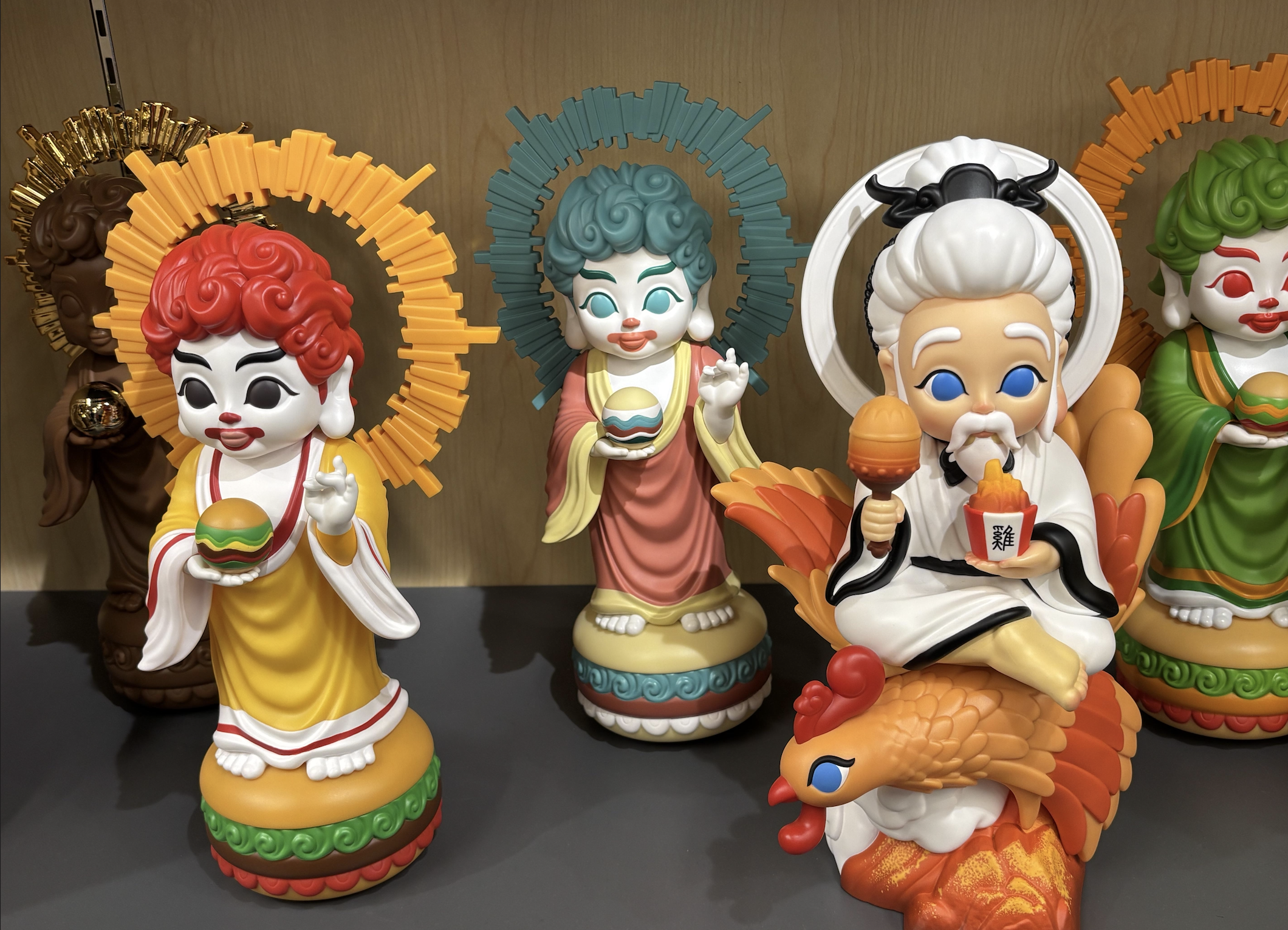 Three colorful figurines stand on a shelf: two with halos and burgers, and one elderly figure with white hair riding a rooster, holding a drink and a scroll. All have cartoon-like features and vibrant colors.