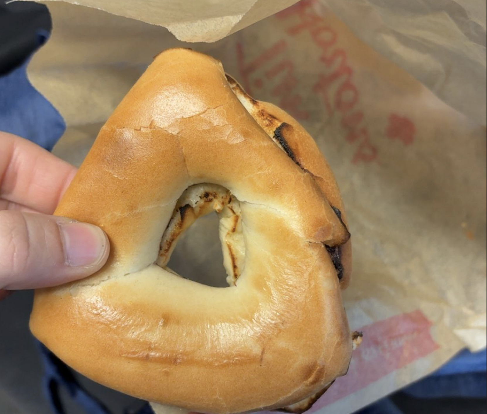 A hand holds a triangular bagel with a heart-shaped hole in the center, above a brown paper wrapper with partial red text.