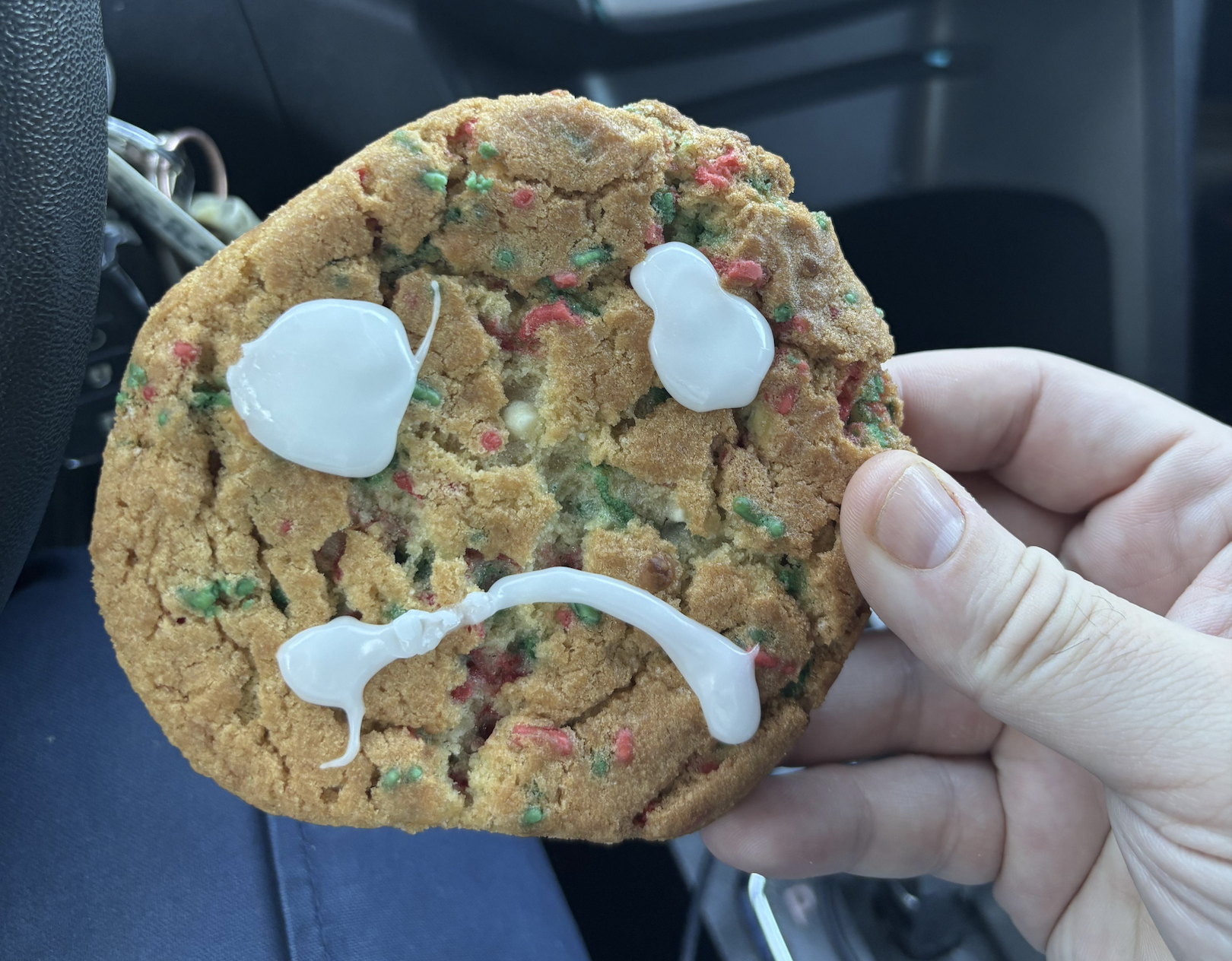 A hand holds a cookie with multicolored sprinkles and white icing arranged to resemble a sad face.