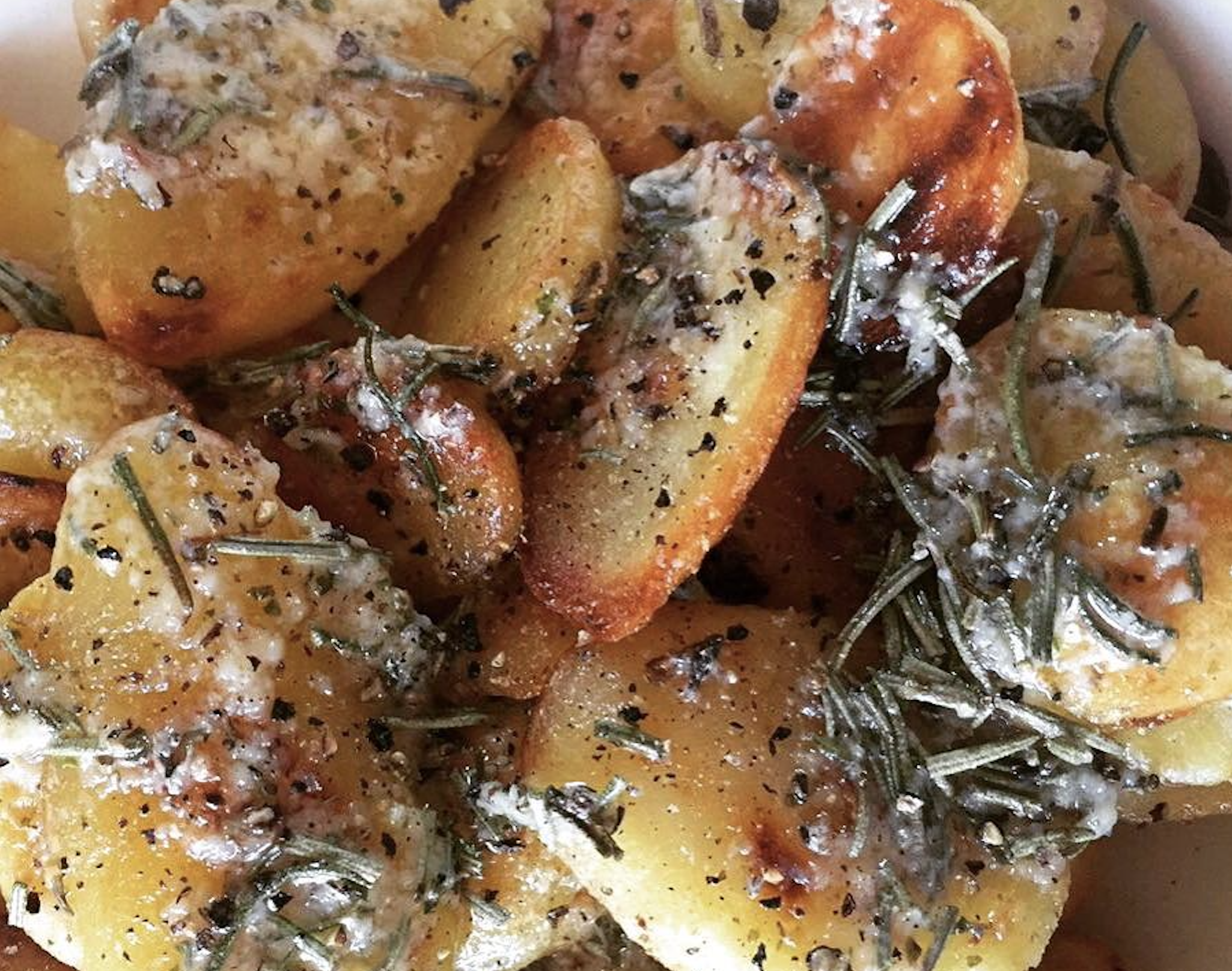 Close-up of roasted potato slices topped with creamy sauce, cracked black pepper, and fresh rosemary. Some potatoes appear golden and crispy, while others are coated with herbs and seasoning.