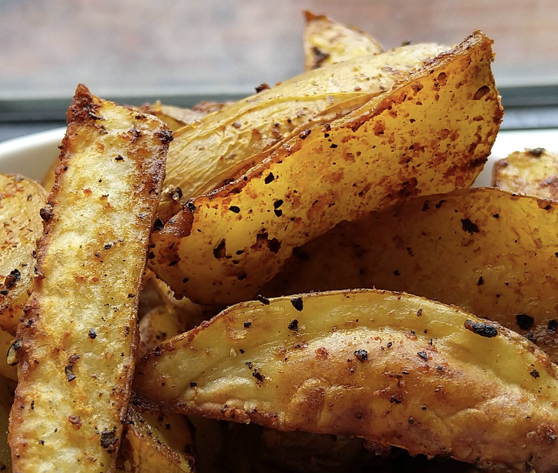 Close-up of crispy, seasoned potato wedges with golden-brown edges, speckled with black pepper and spices, piled together in a bowl.