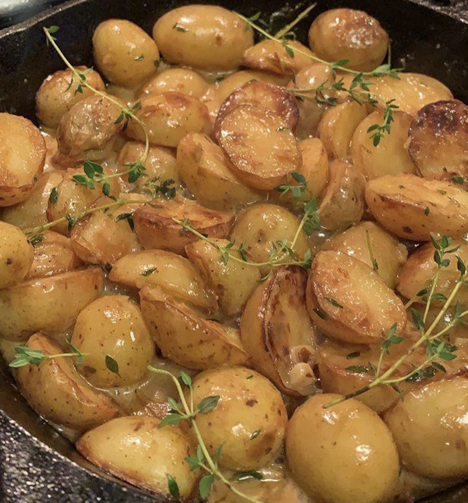 Golden roasted baby potatoes with fresh thyme sprigs, glistening with oil and seasonings, are cooked in a black skillet.