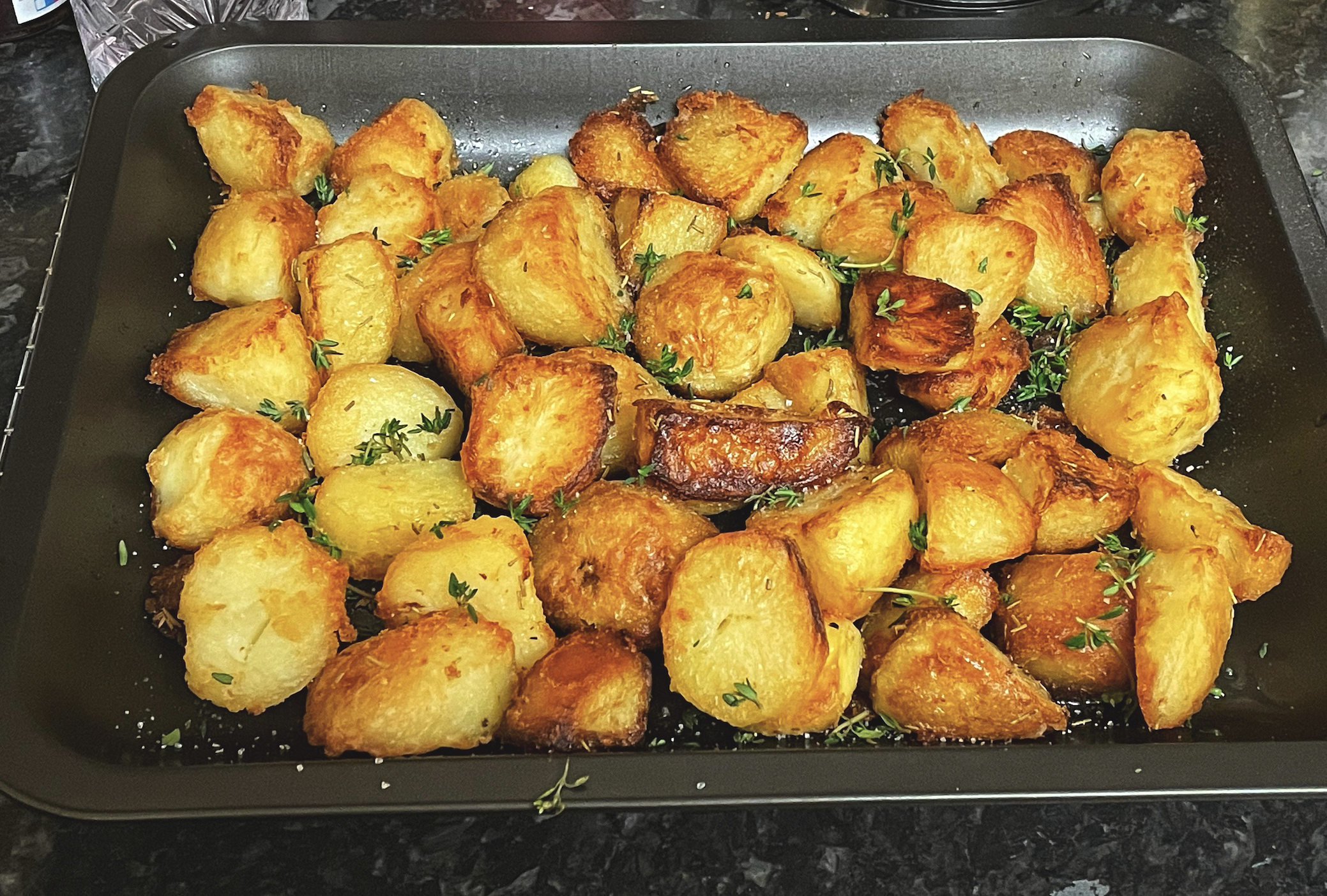A baking tray filled with golden, crispy roasted potatoes garnished with fresh herbs, sitting on a dark countertop.