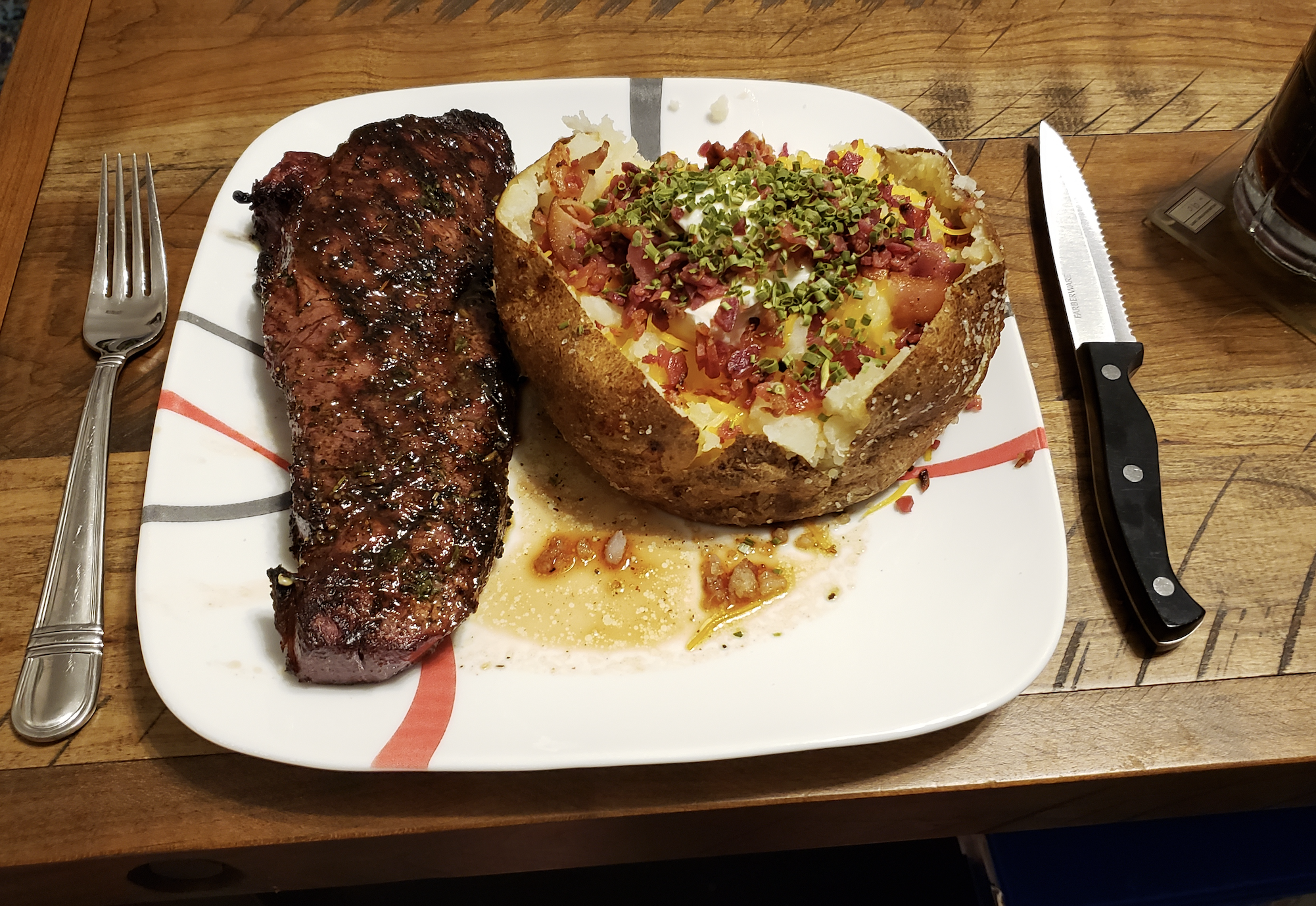 Grilled steak and a loaded baked potato topped with sour cream, bacon bits, cheese, and chives on a white plate with a steak knife on a wooden table.