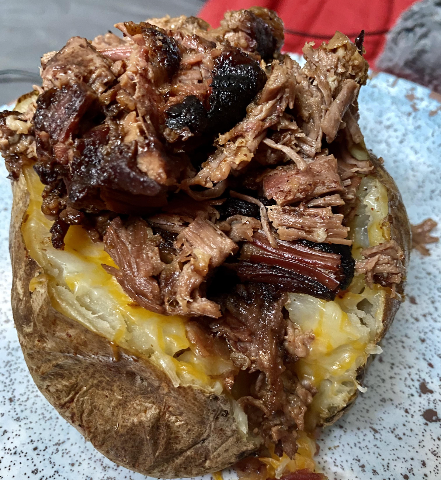 A baked potato topped with shredded, seasoned beef and melted cheese on a white speckled plate.