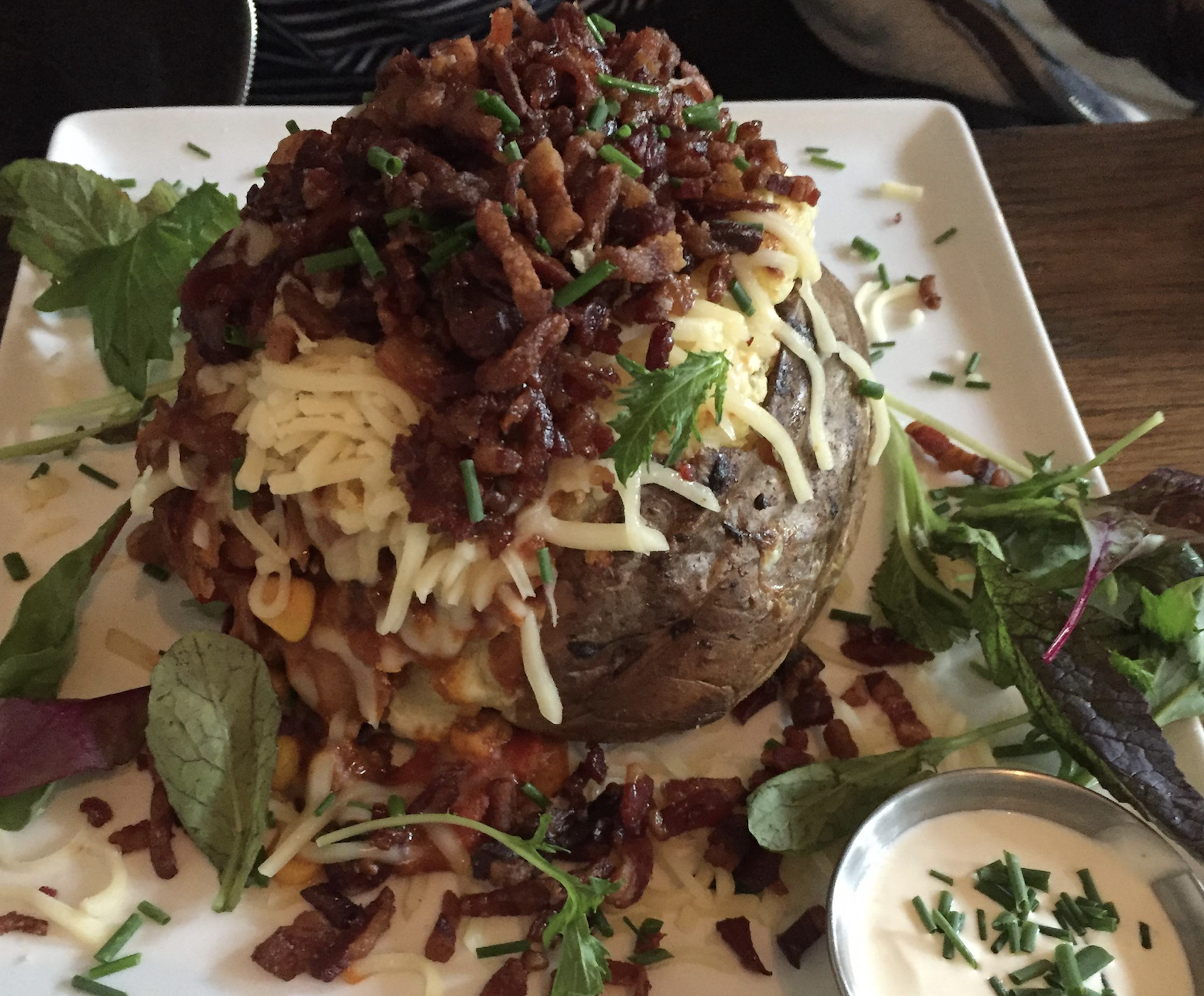 A baked potato topped with shredded cheese, crispy bacon bits, and chives, served on a plate with mixed greens and a small cup of sour cream.