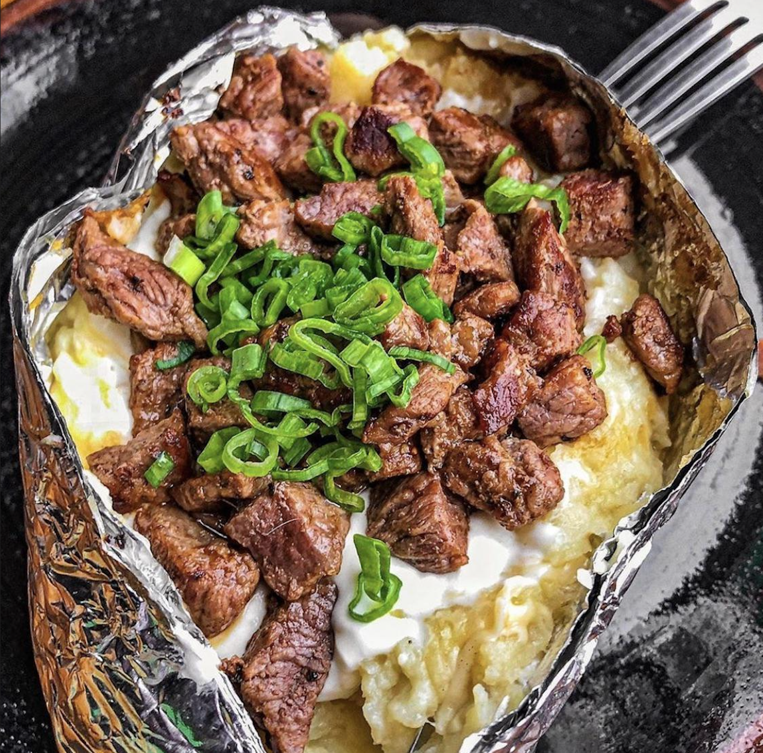 A baked potato cut open and topped with sour cream, grilled steak pieces, and chopped green onions, served on a black plate with a fork visible in the background.