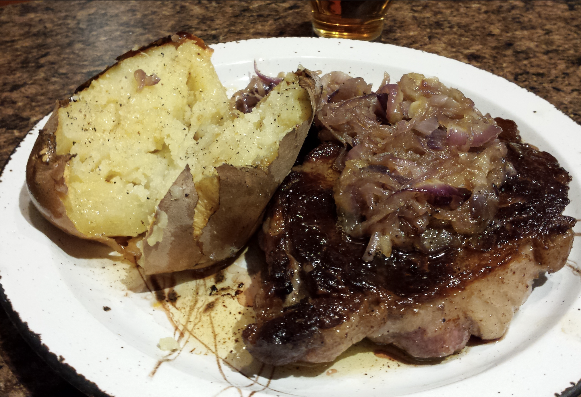 A baked potato split open with butter and pepper sits beside a grilled steak topped with sautéed onions on a white plate, placed on a brown countertop.