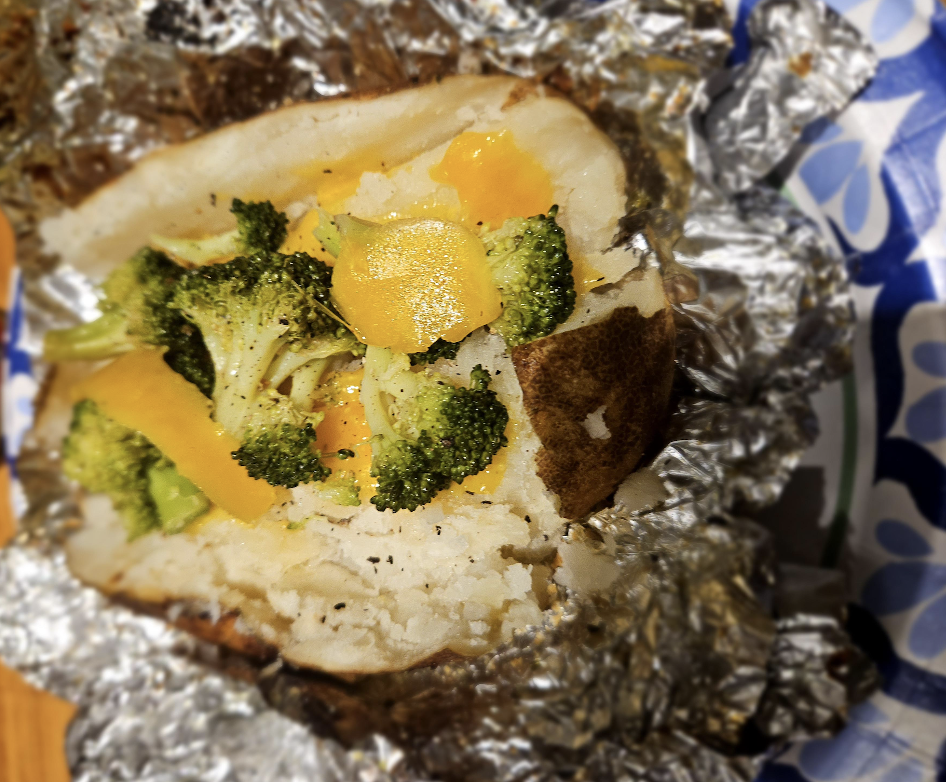 A baked potato wrapped in foil, topped with broccoli florets and melted cheddar cheese, sits on a patterned plate.