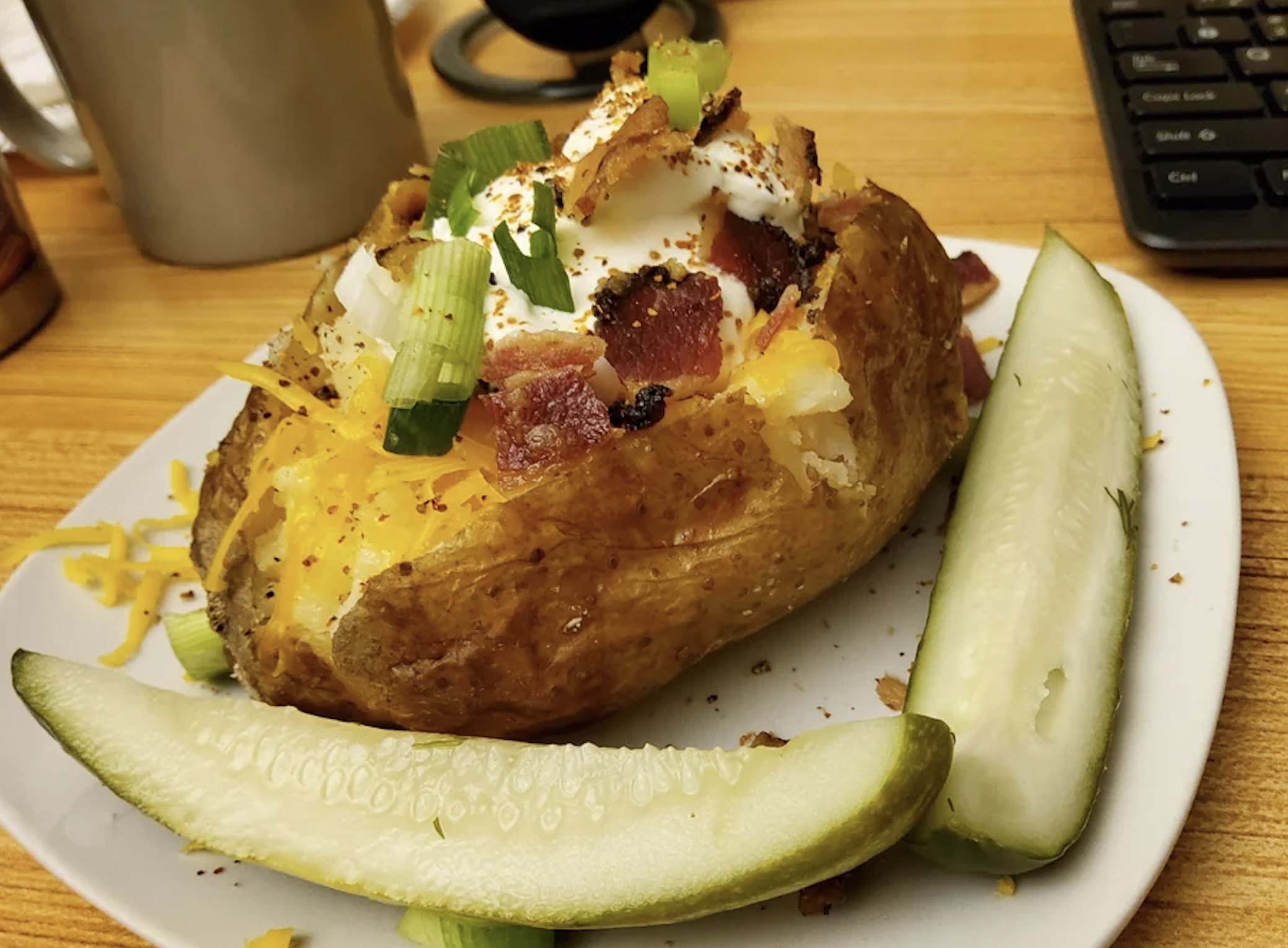 A baked potato topped with sour cream, bacon, cheese, and green onions sits on a plate with two pickle spears. Part of a keyboard and mug are visible in the background.