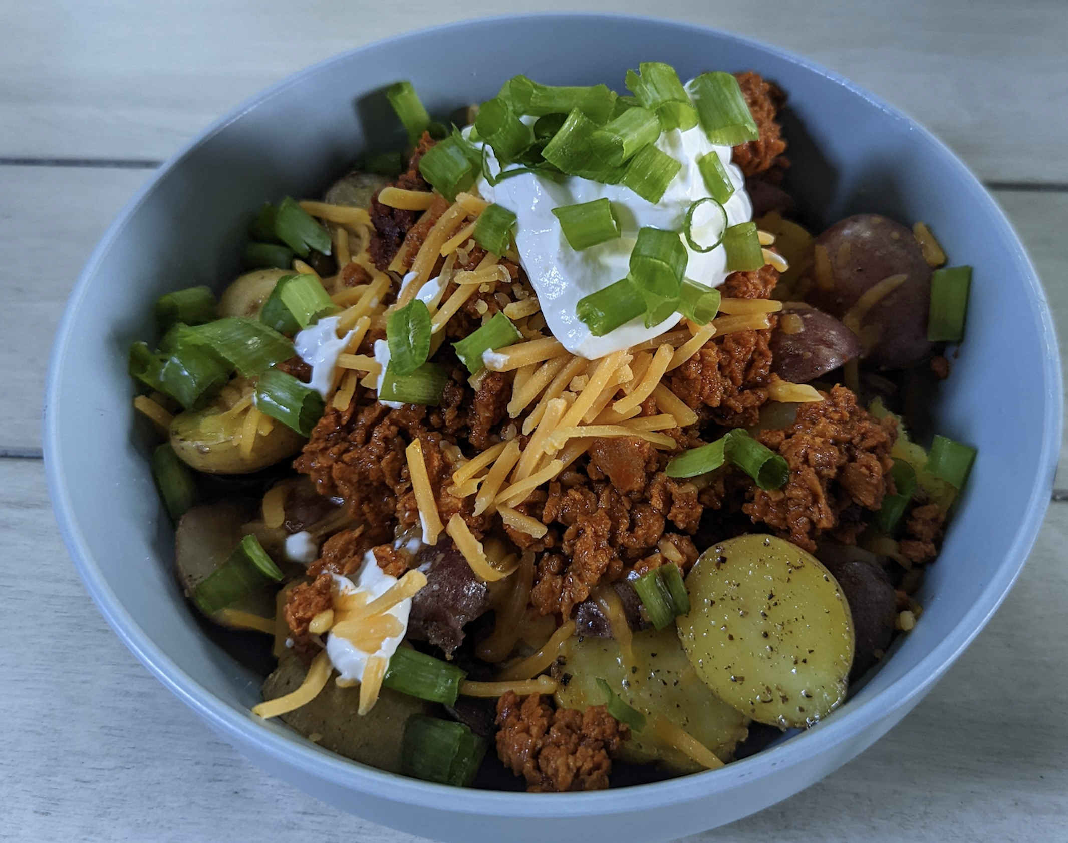 A bowl of sliced potatoes topped with seasoned ground meat, shredded cheddar cheese, sour cream, and chopped green onions, served on a light wooden surface.