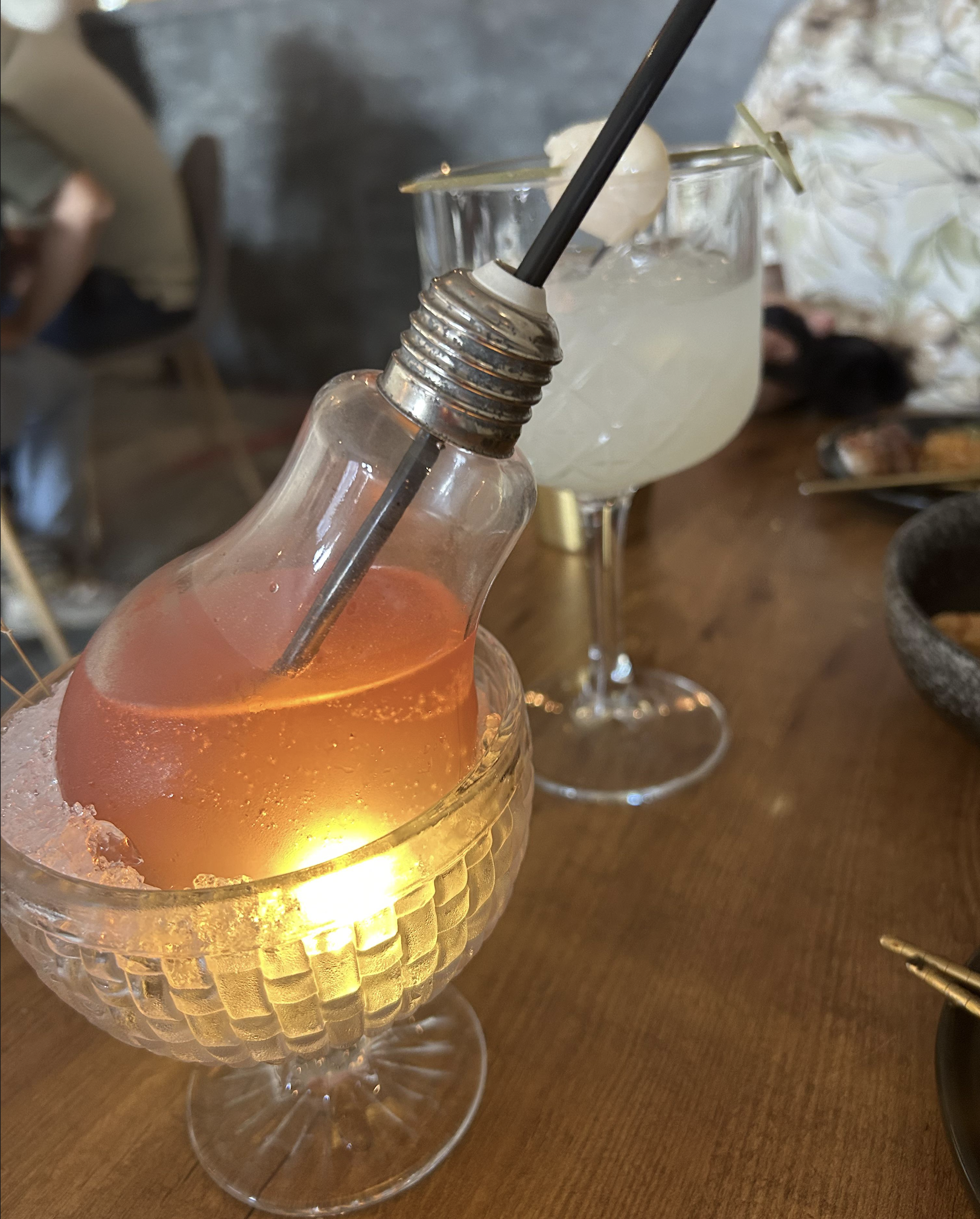 A cocktail served in a lightbulb-shaped glass with a straw sits in a crystal bowl, beside another cocktail in a wine glass with a lychee garnish on a wooden table.