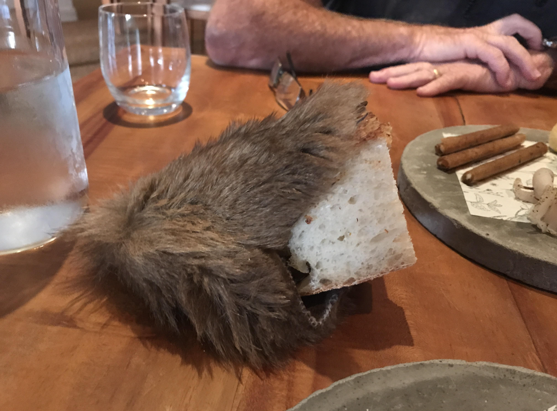 A piece of bread is partly covered by brown faux fur, resting on a wooden table with glassware and plates nearby. A person’s arms are visible in the background.