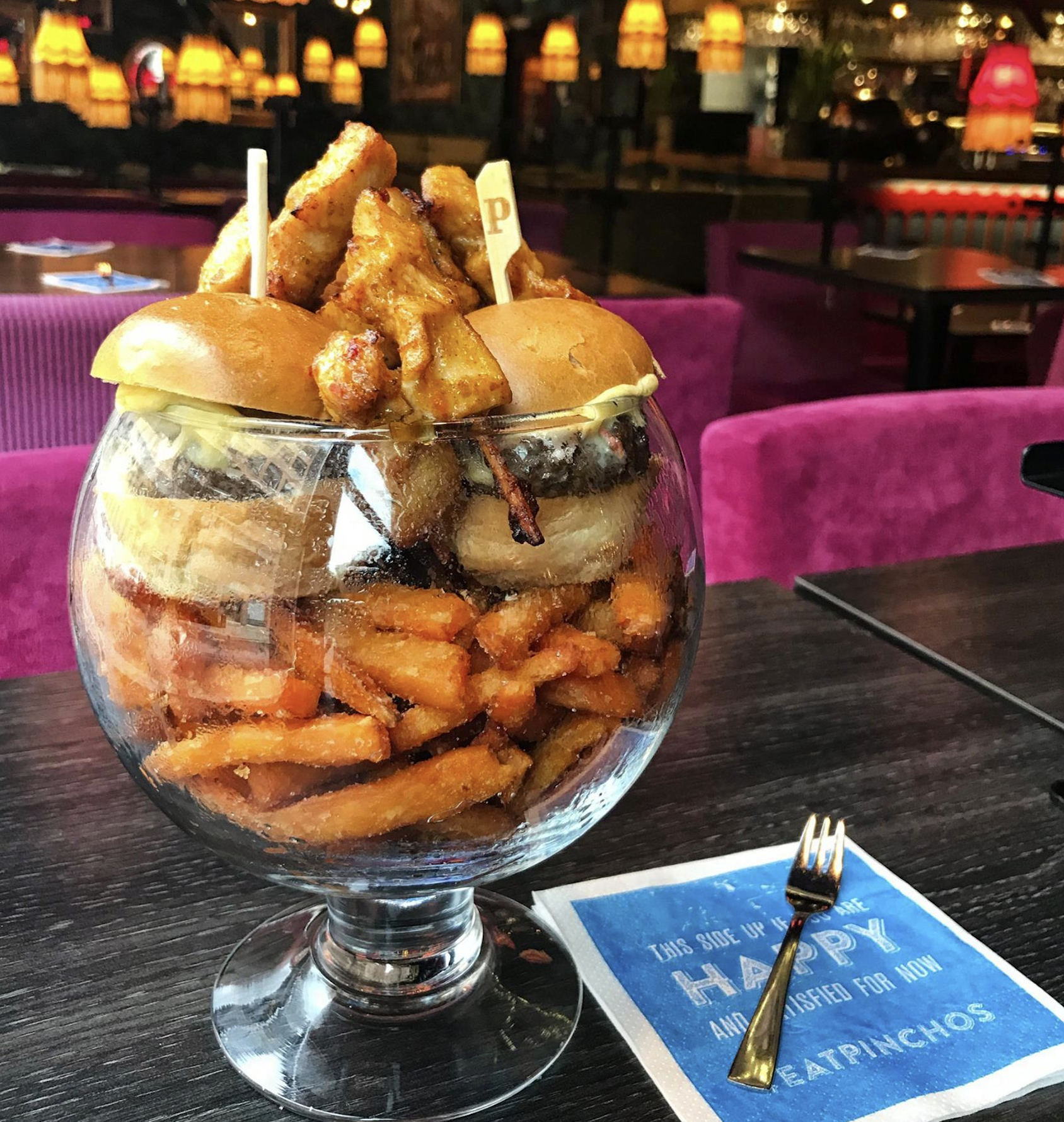 A large glass bowl filled with sweet potato fries and topped with two burgers and grilled skewers, sitting on a dark table in a restaurant with purple chairs and warm lighting.