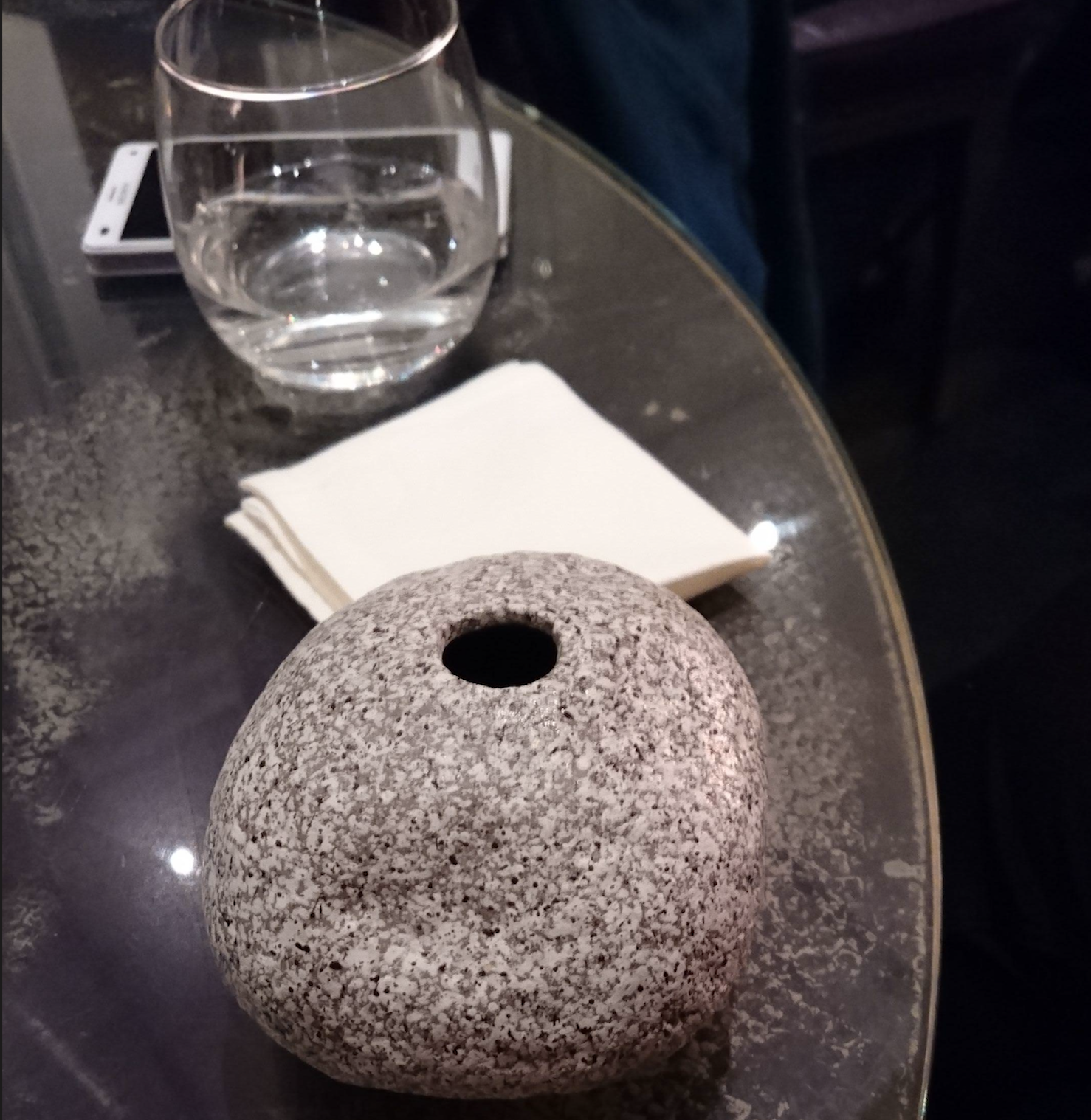 A round, gray stone with a hole sits on a glass table next to a stack of white napkins and a clear water glass. Part of a smartphone is visible under the table.