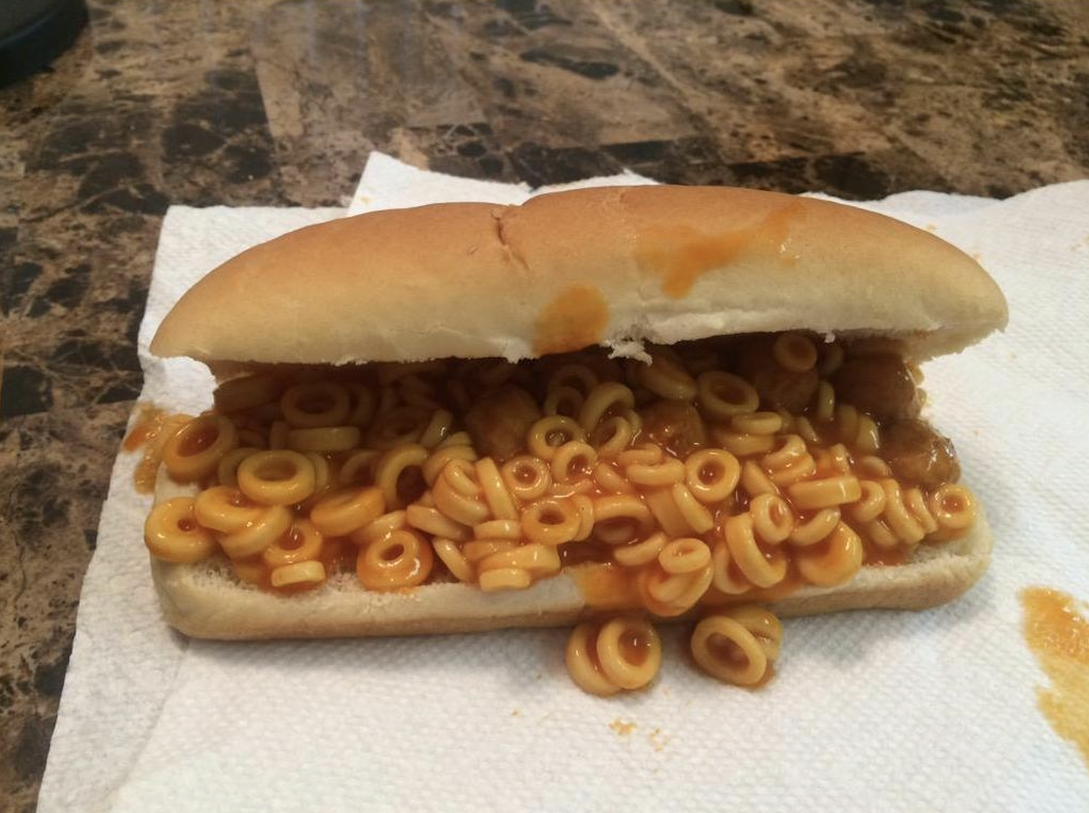 A hot dog bun filled with canned spaghetti rings in tomato sauce sits on a white napkin on a marble table.