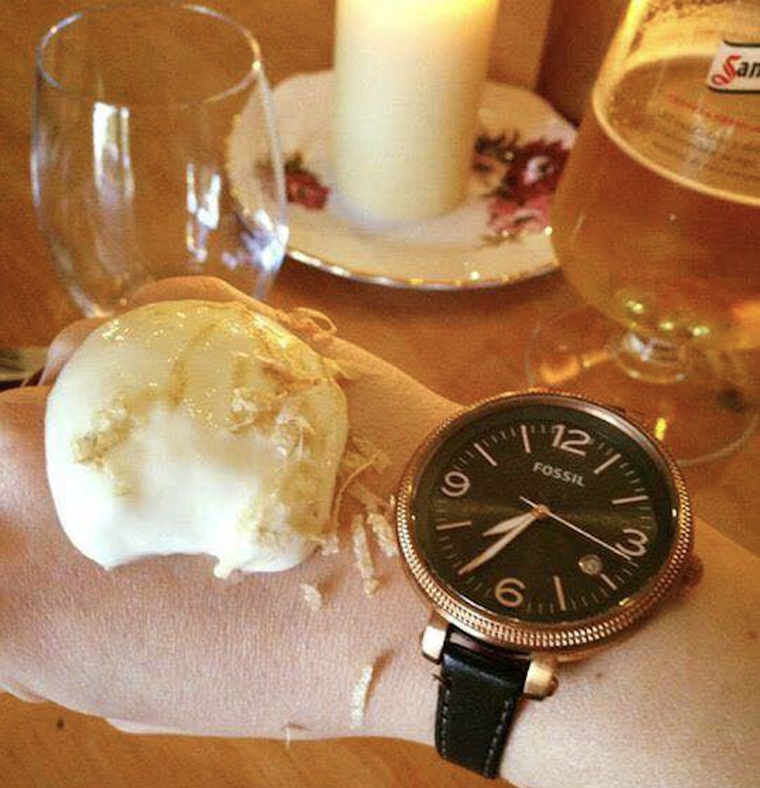A person wearing a wristwatch holds a scoop of melting ice cream on the back of their hand. A candle, glass, and beer are on the table.