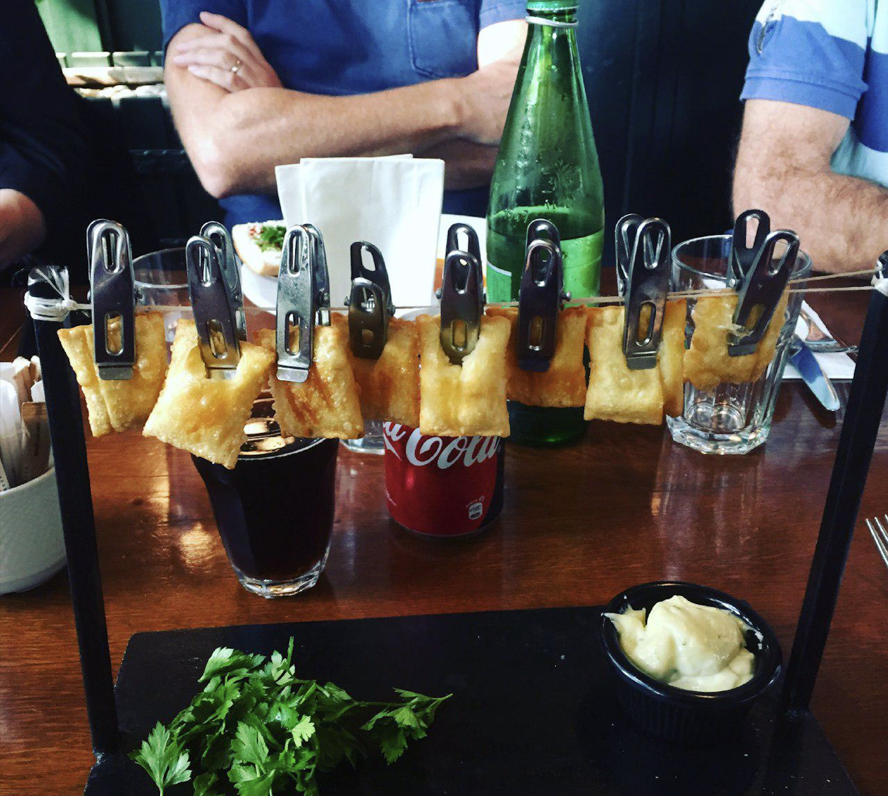 Several crispy snacks are clipped to a hanging rod above a tray with herbs and a sauce; drinks and glasses are on the table, and people sit in the background.