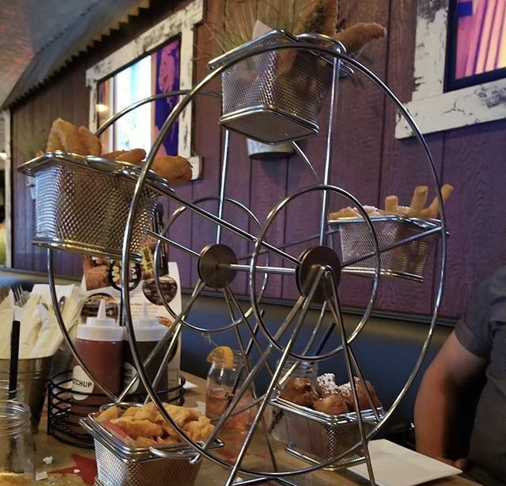 A metal Ferris wheel-shaped server holds baskets filled with fried appetizers on a restaurant table. Drinks, sauces, and menus are also visible, with part of a person sitting at the table.