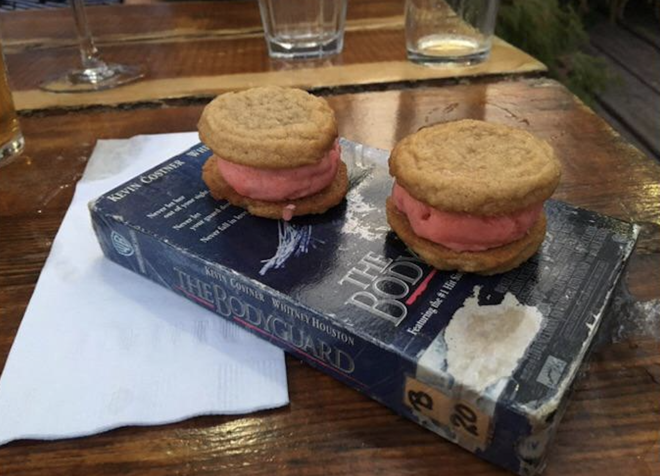 Two ice cream sandwiches made with cookies and pink ice cream rest on a worn VHS tape of “The Bodyguard.” Nearby are a napkin, a glass, and other glasses on a wooden outdoor table.
