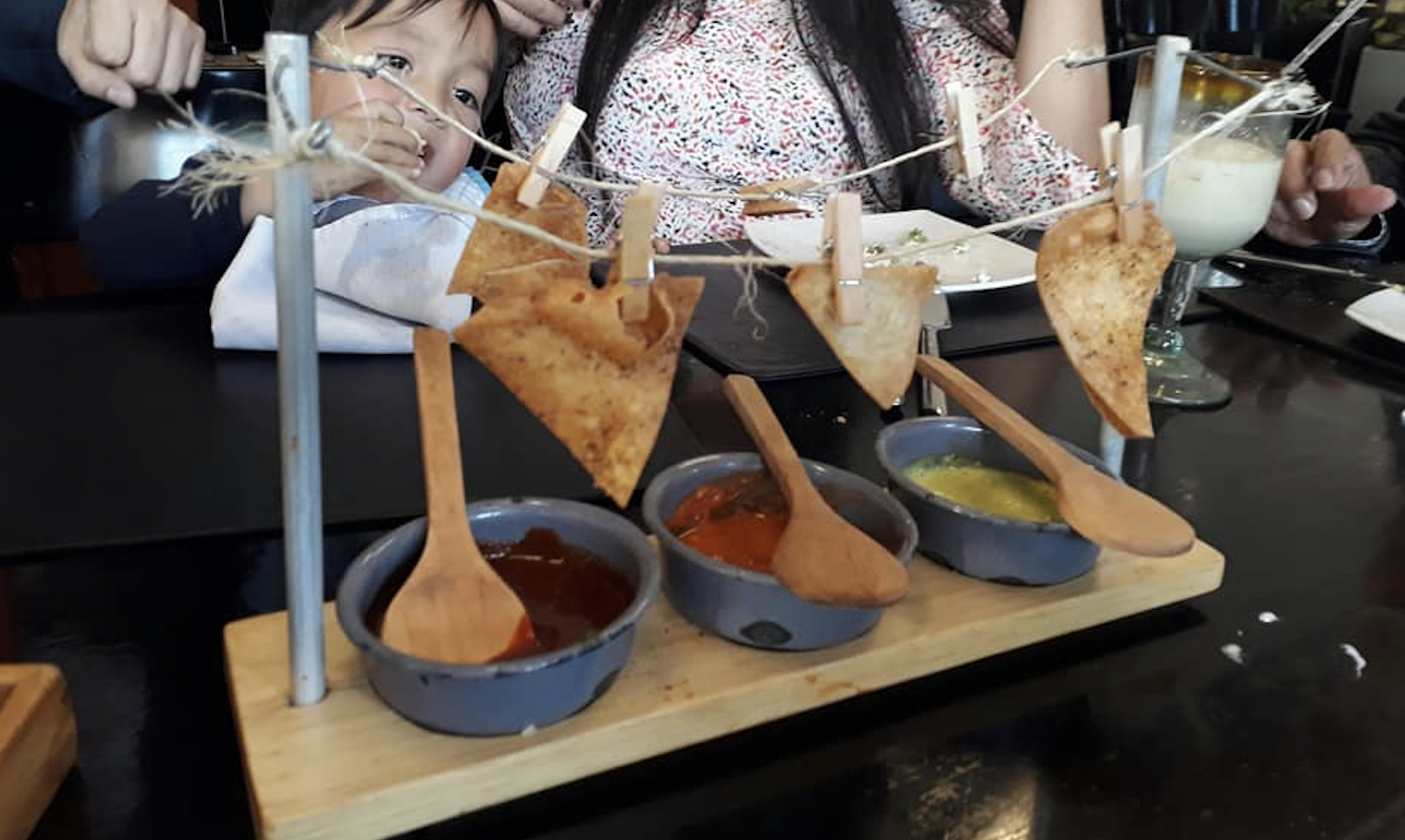 Three tortilla chips hang on strings above three bowls of dips with wooden spoons, served on a wooden tray. A woman and child are partially visible in the background at a dining table.