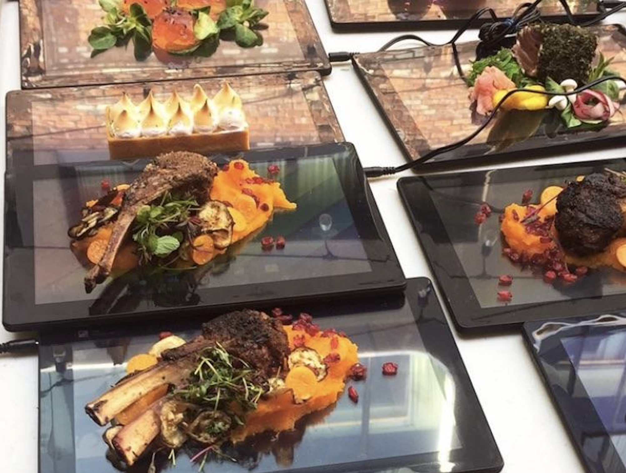 Plates of gourmet food are served on top of digital tablets, displaying brick patterns, with dishes including lamb chops, dessert, and assorted vegetables.