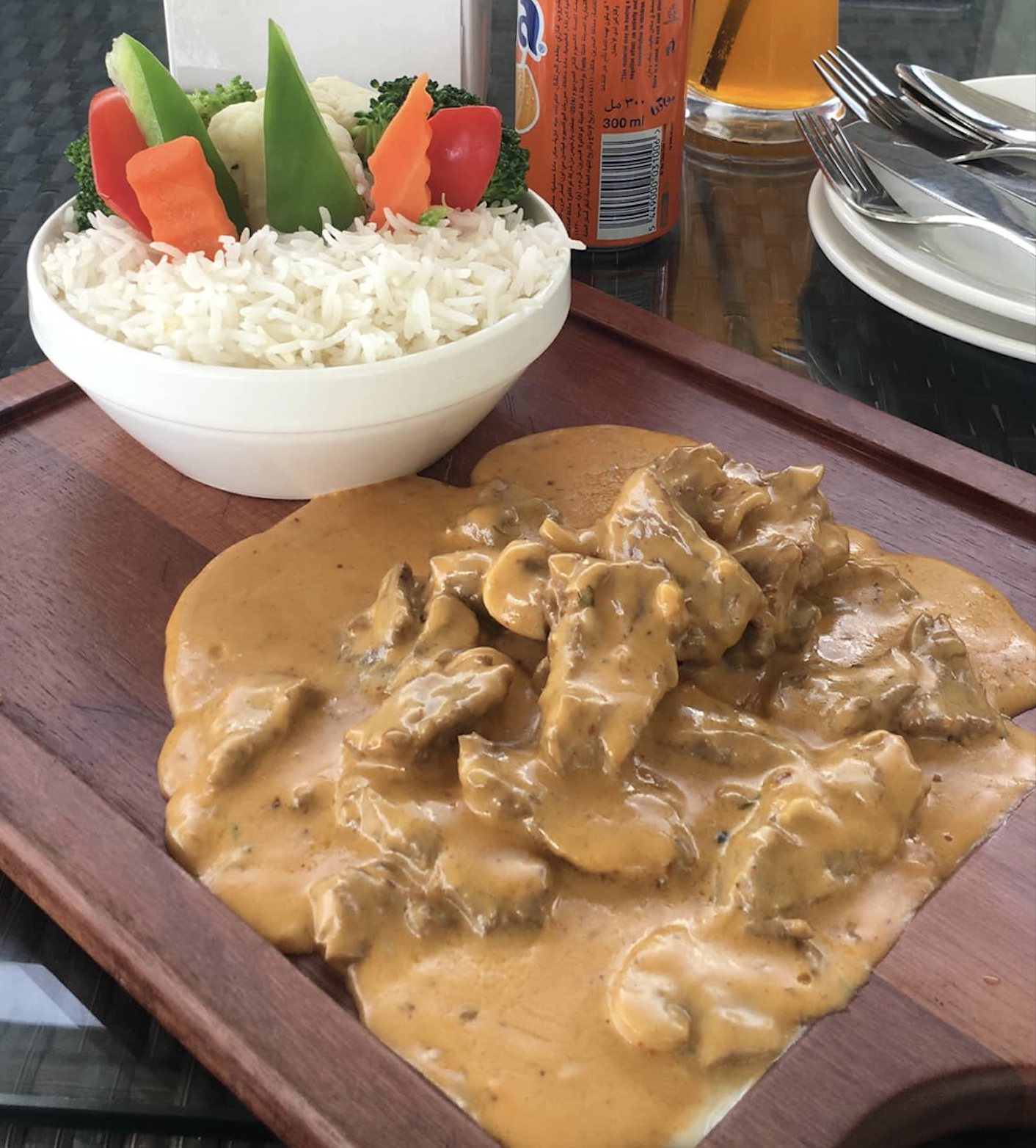 A wooden tray with creamy beef stroganoff and a bowl of white rice topped with colorful steamed vegetables. Plates, cutlery, a soda can, and a glass are in the background.