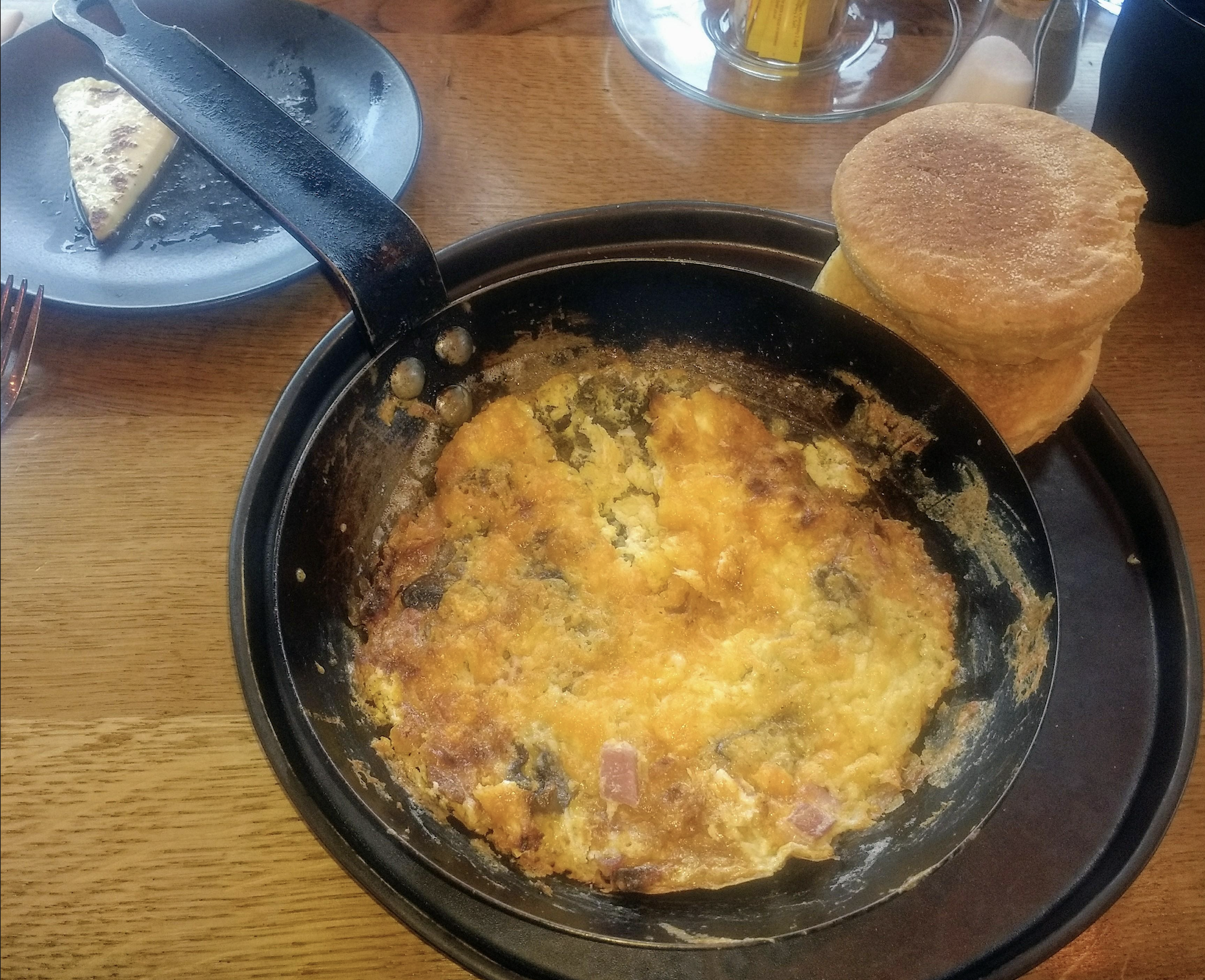 A skillet with a baked omelette containing ham and vegetables sits on a table next to two English muffins and a plate with a small slice of toasted bread.