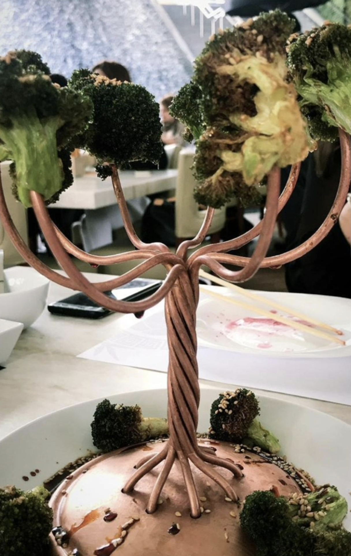 A creative dish features broccoli “trees” on twisted metal branches, resembling a tree, atop a dome with sauce and sesame seeds, served in a restaurant.