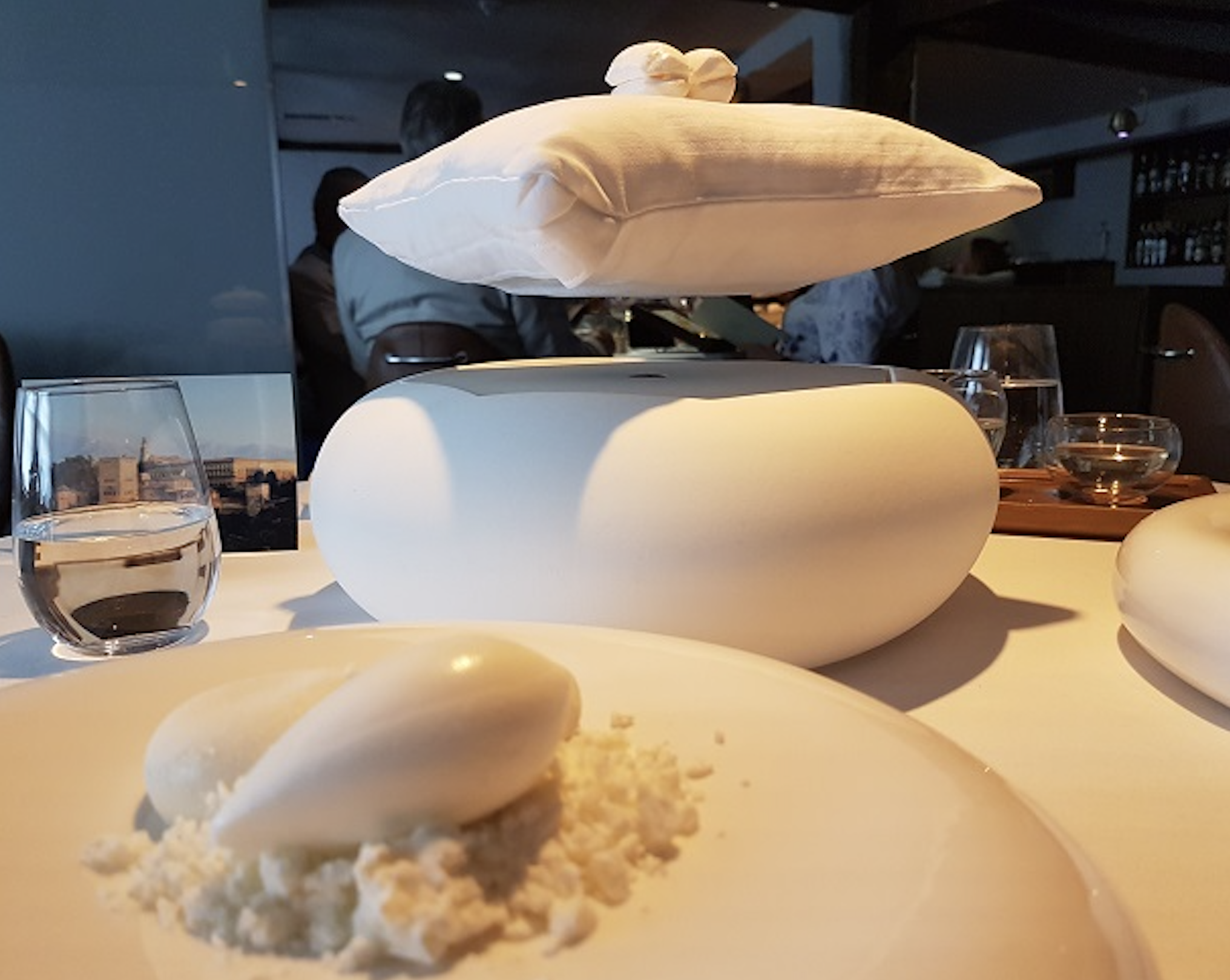 A white pillow floats above a rounded base on a dining table, with an elegant dessert and glass of water in the foreground. The setting appears modern and upscale.