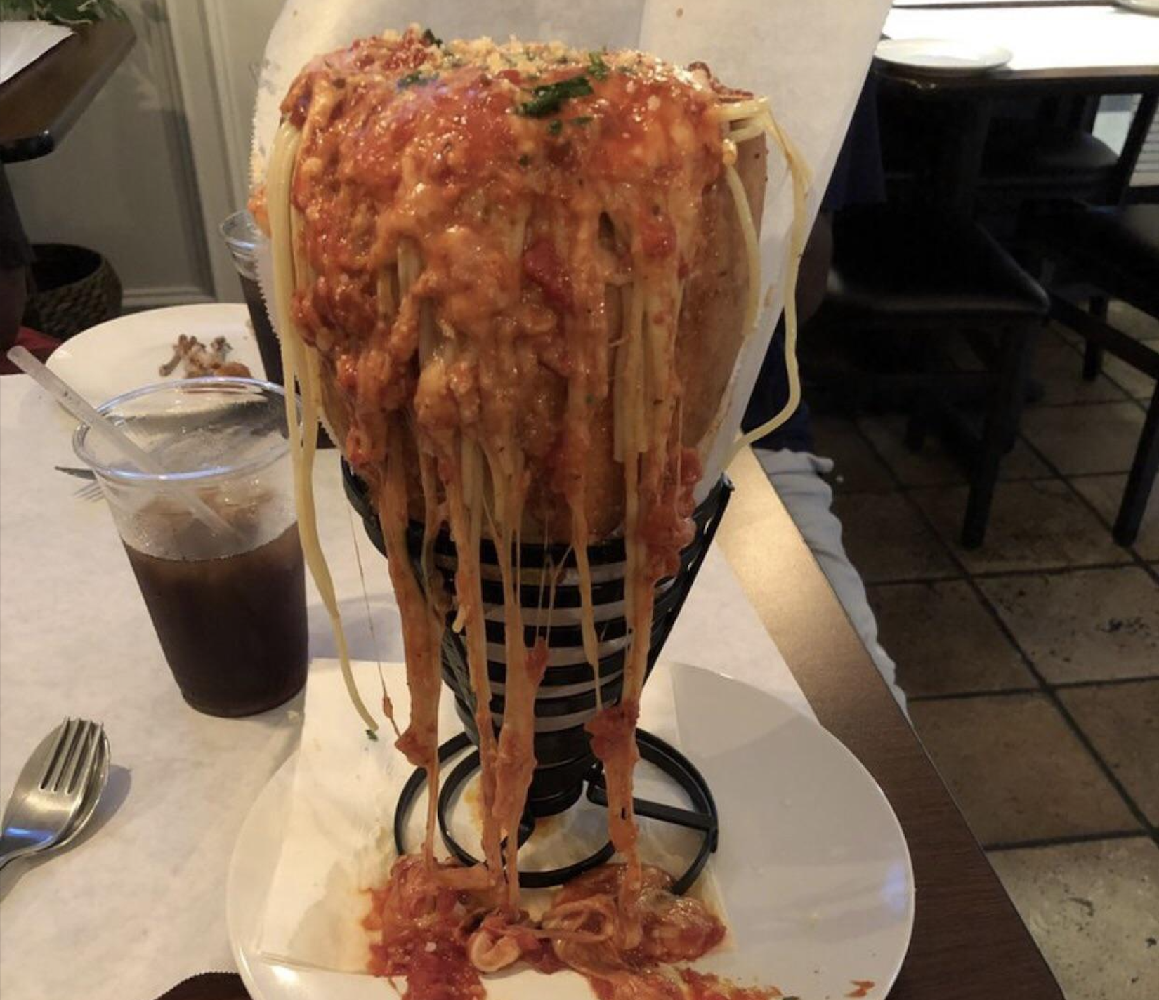 A large serving of spaghetti with red sauce overflows from a vertical cone-shaped holder, with noodles and sauce dripping down onto a plate below. Iced drink, fork, and napkin are on the table.