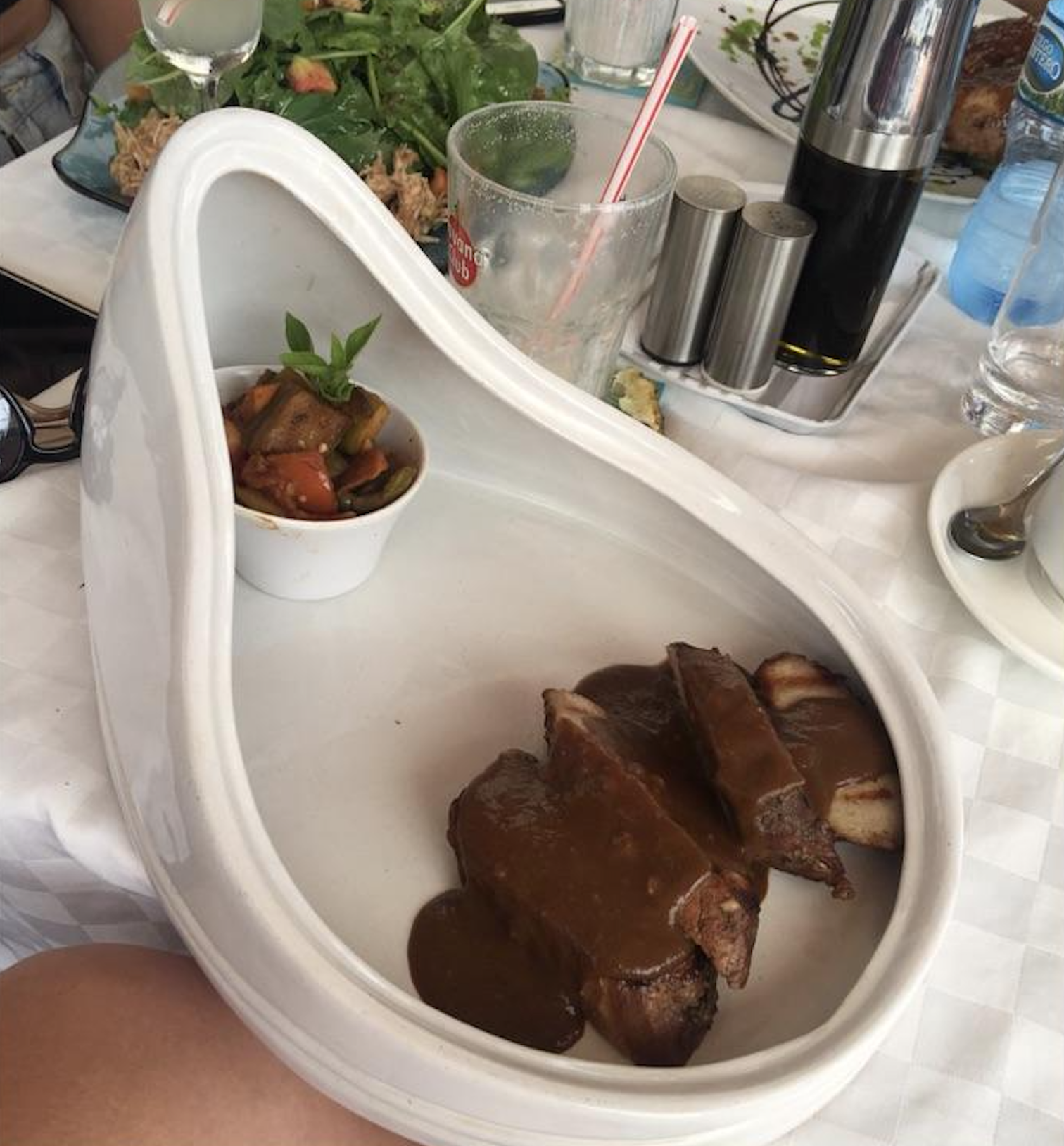 Slices of meat with brown sauce and a side of vegetables served in a dish shaped like a urinal, on a table with salad, drinks, and condiments in the background.