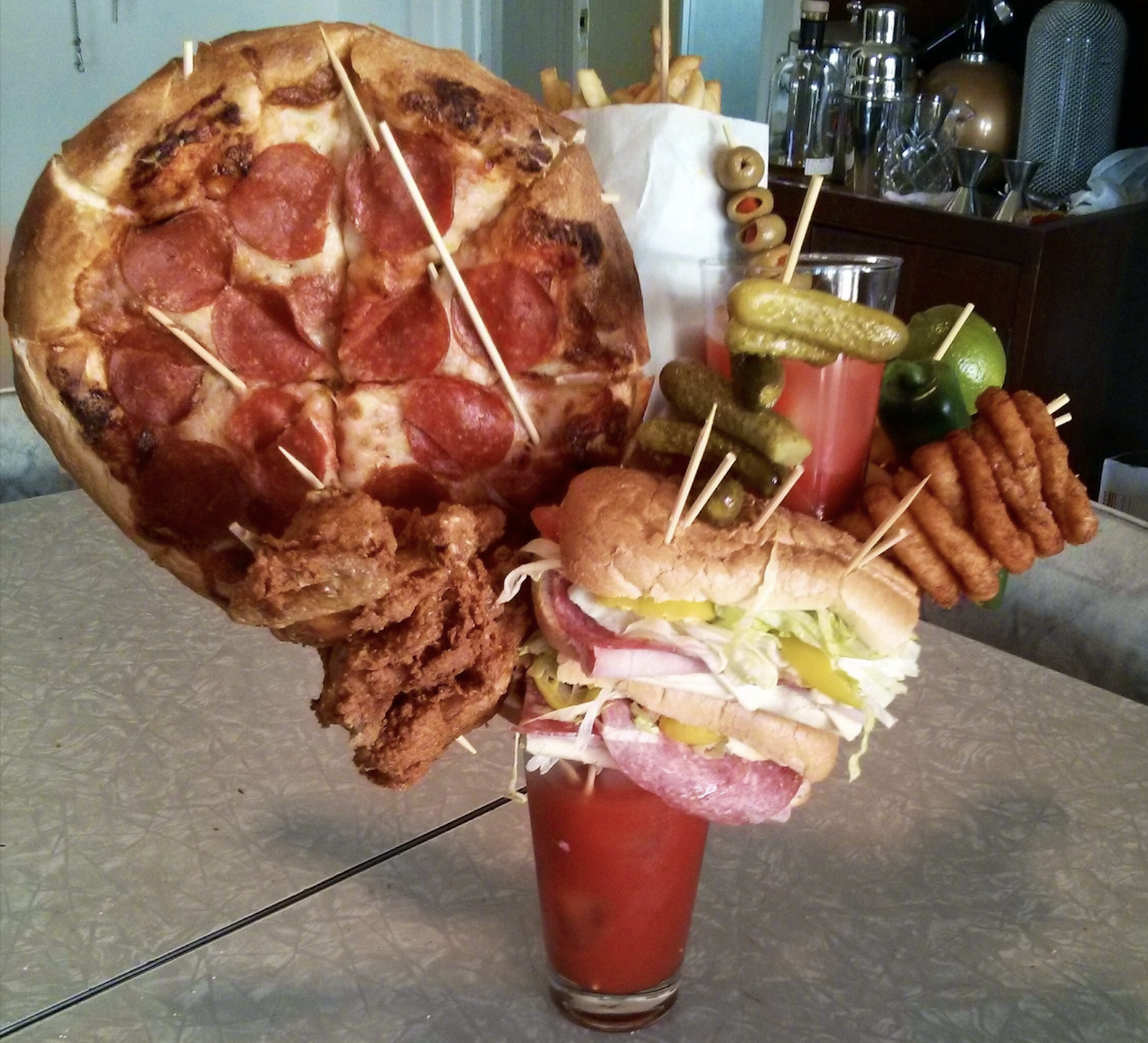 A tall glass of Bloody Mary is topped with skewers holding fried chicken, a sandwich, curly fries, pickles, olives, limes, and a large slice of pepperoni pizza, with a bag of fries in the background.