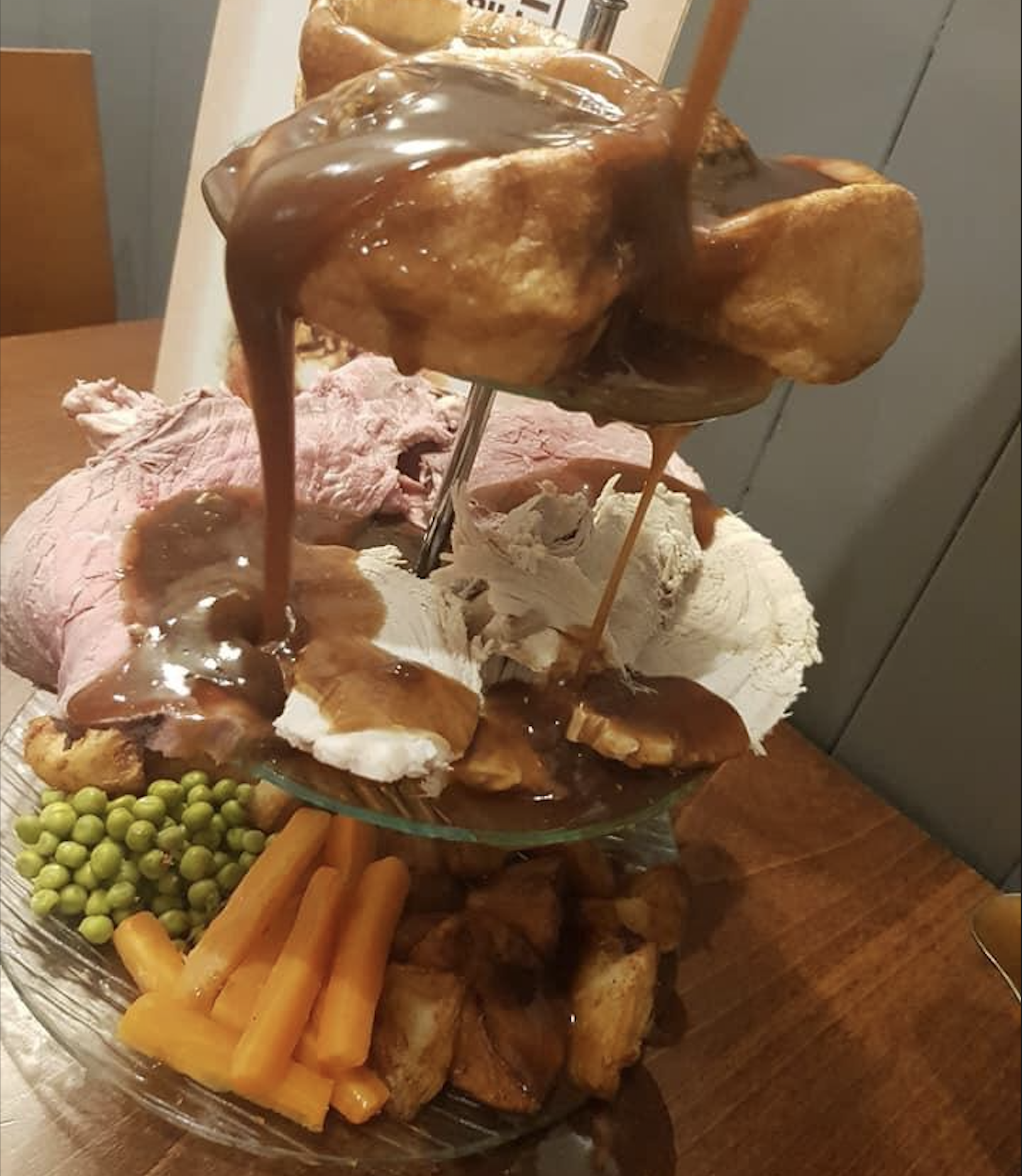 A tiered platter with roast beef, turkey, Yorkshire puddings, carrots, peas, roast potatoes, and gravy being poured over the top, set on a wooden table.