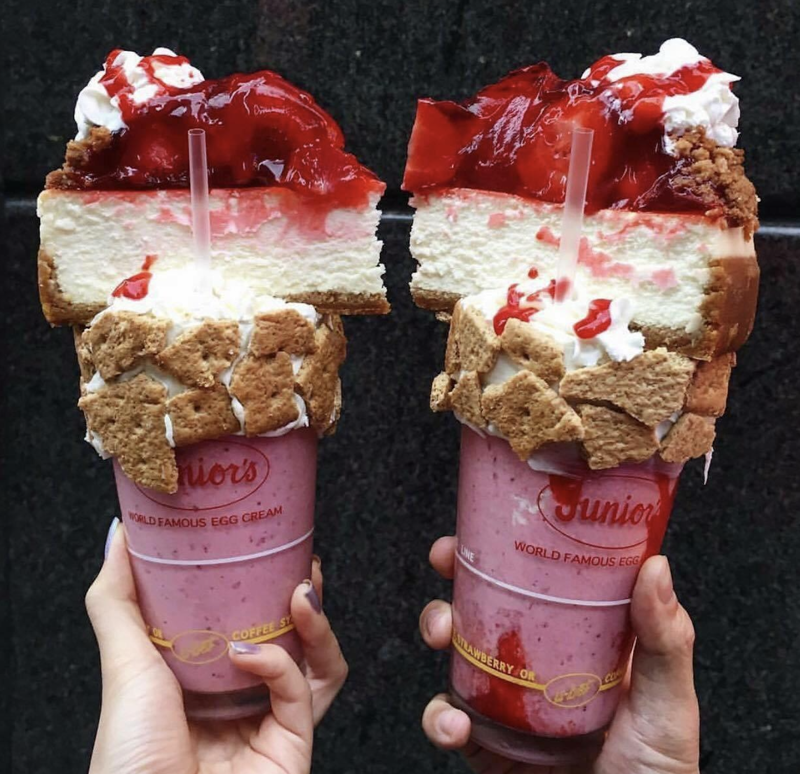 Two hands hold milkshakes topped with whipped cream, strawberry sauce, crumbled graham crackers, and a large slice of strawberry cheesecake in clear Junior’s cups against a dark background.